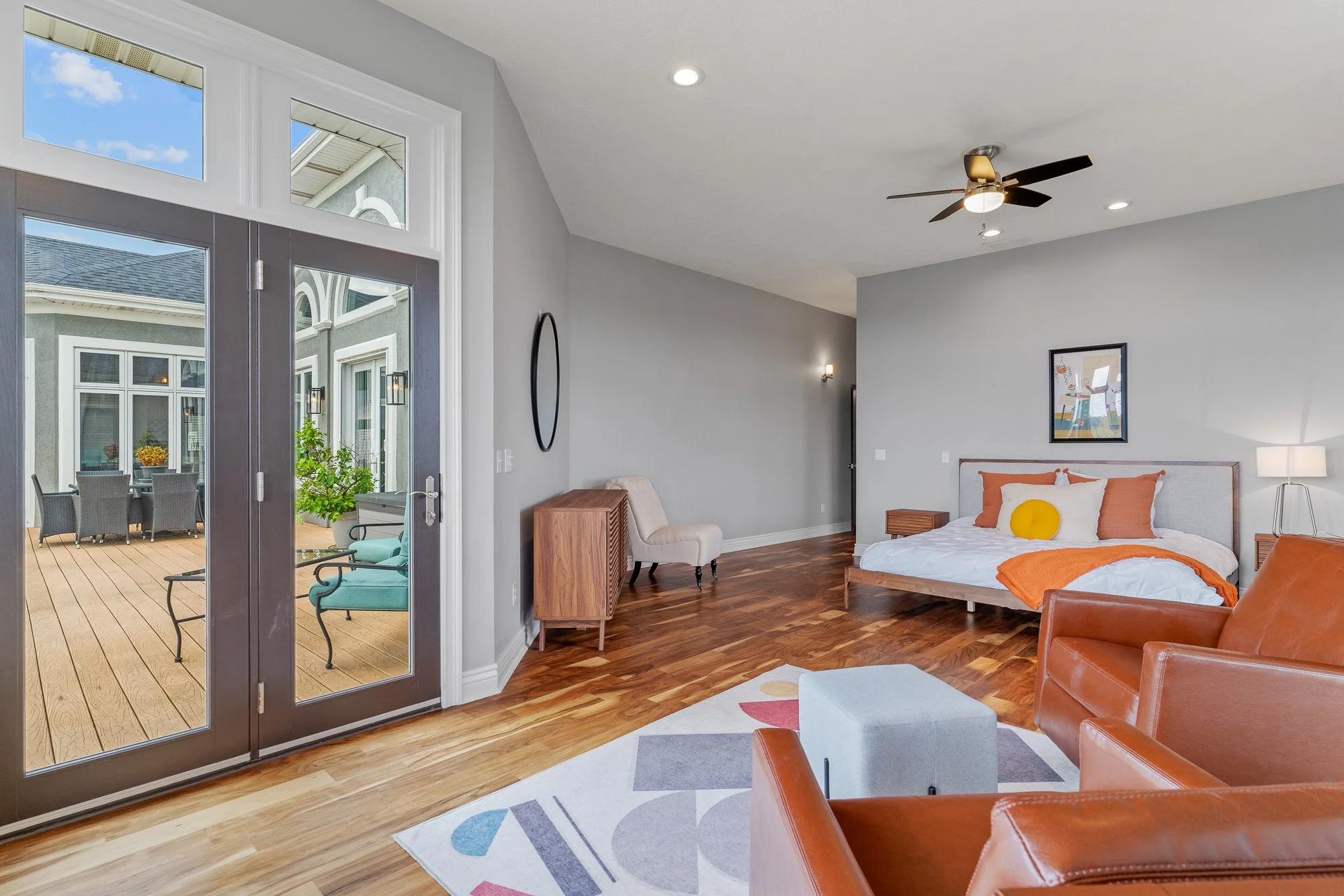 A bedroom with hardwood floors, a bed with white and orange bedding, and pillows, a framed picture on the wall, a ceiling fan, a small white ottoman, and a glass door leading to a balcony with outdoor table and chairs.