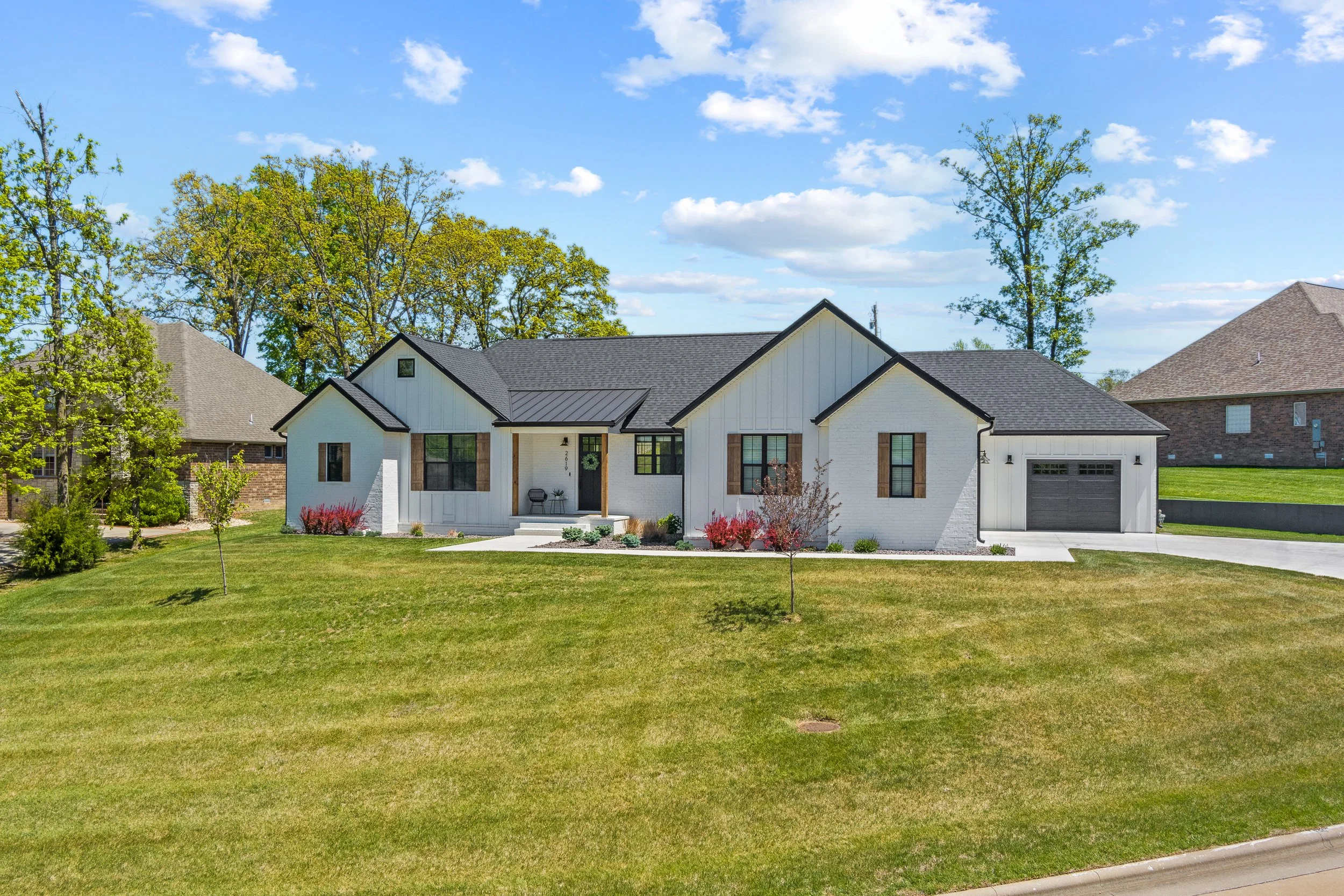 A modern white house with a gray roof, front porch, and attached garage, situated on a well-manicured lawn with small trees and bushes, under a partly cloudy blue sky.