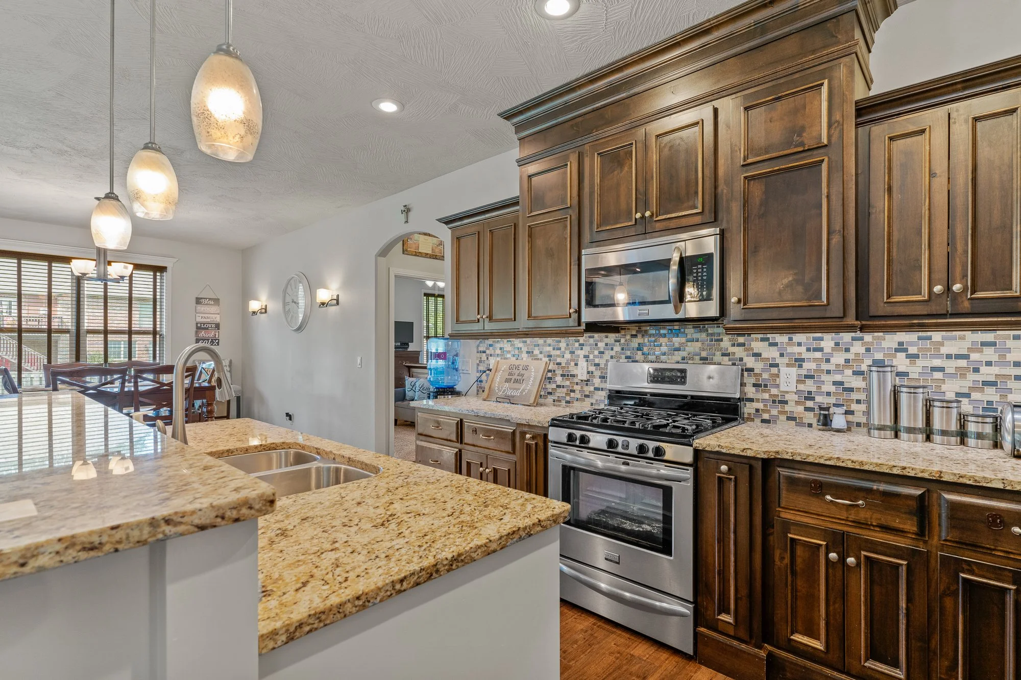 Kitchen with wooden cabinets, granite countertops, stainless steel oven and microwave, tiled backsplash, and a countertop with a sink and water dispenser.