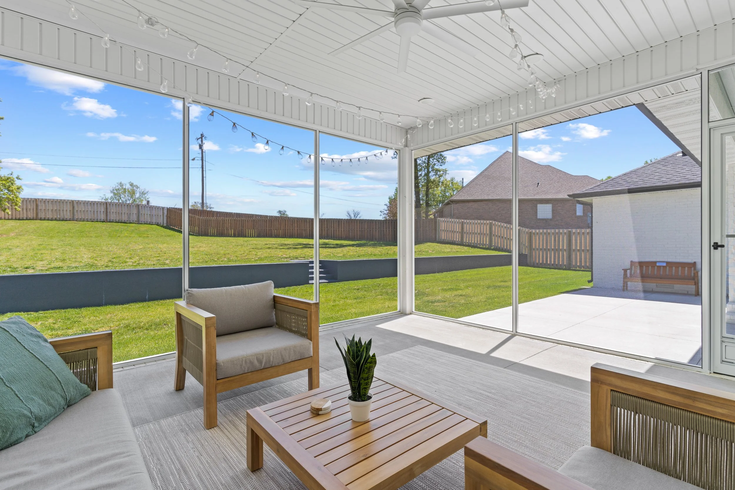 A screened-in patio with outdoor furniture including a cushioned sofa, armchairs, and a wooden coffee table with a small potted plant. String lights hang from the ceiling. The backyard has a green lawn, a wooden fence, and neighboring houses visible 