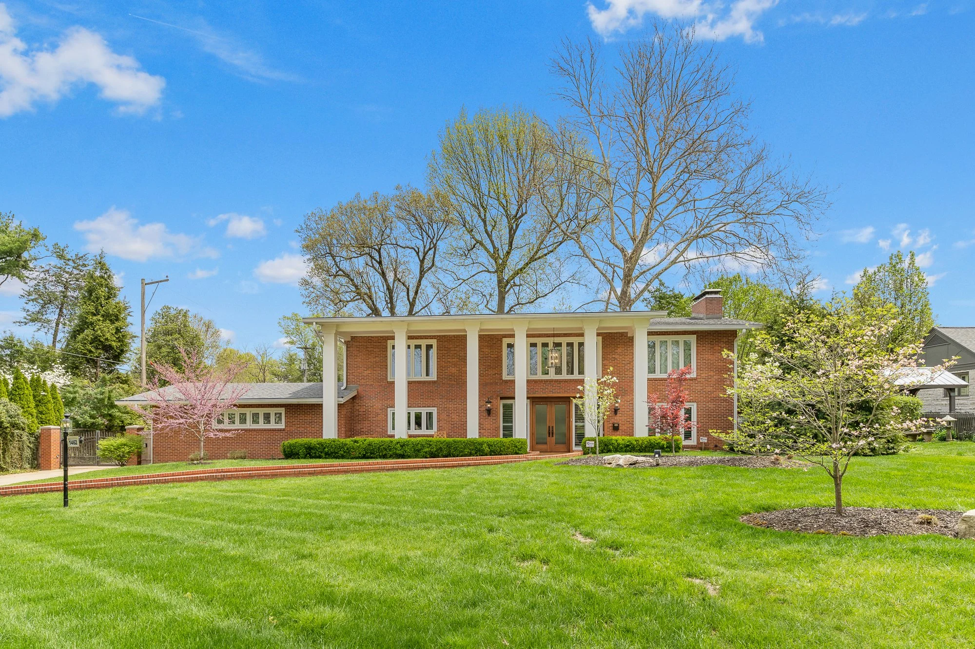 Front view of a brick house with a large porch, surrounded by a well-manicured lawn and various trees including a deciduous tree with budding leaves and pink flowering trees.
