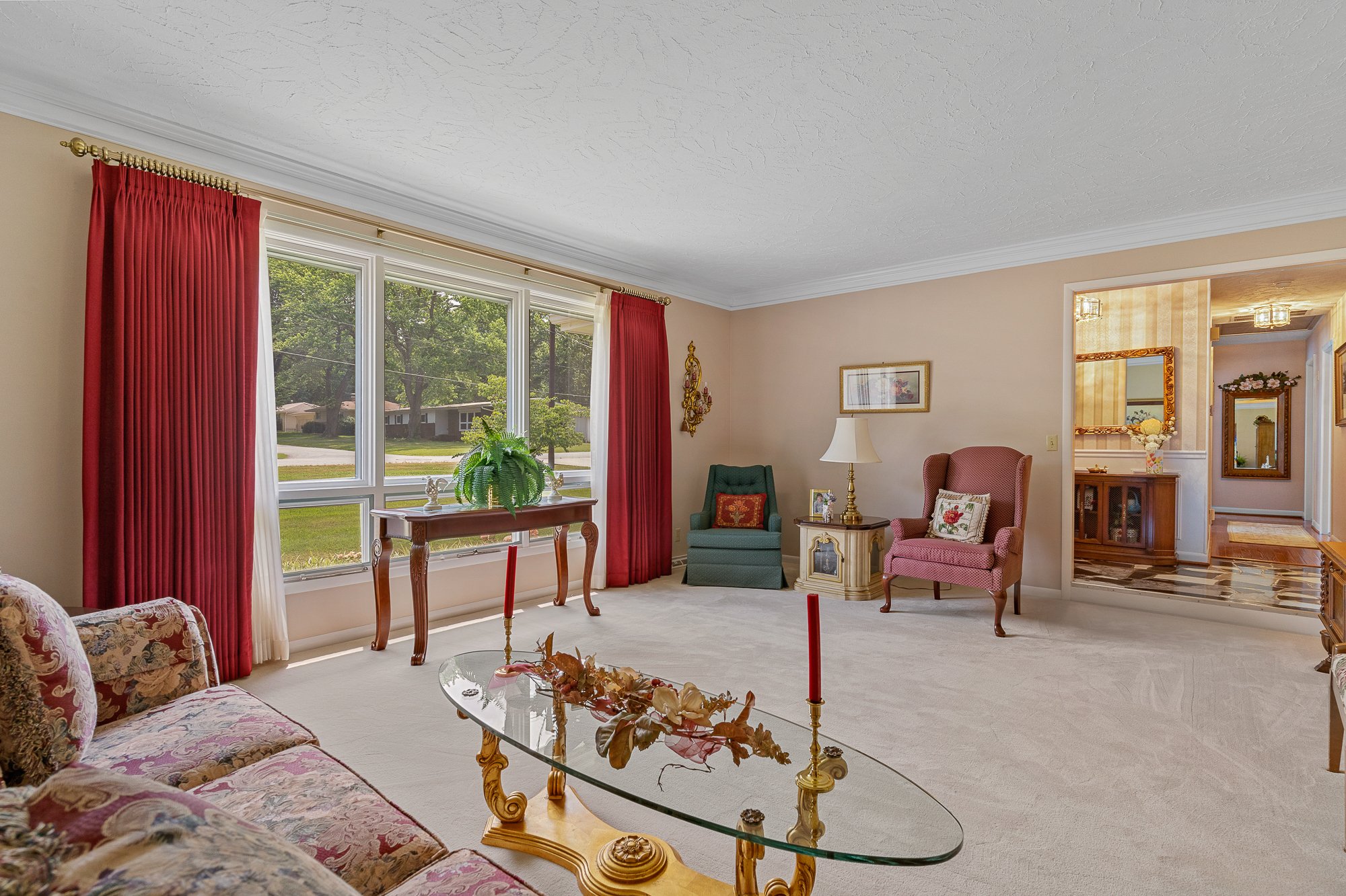 Living room with large window, red curtains, floral upholstered sofa, glass coffee table with decorative leaves and candles, green and pink armchairs, side table with lamp, wall art, and hallway leading to another room with mirror and floral decorati