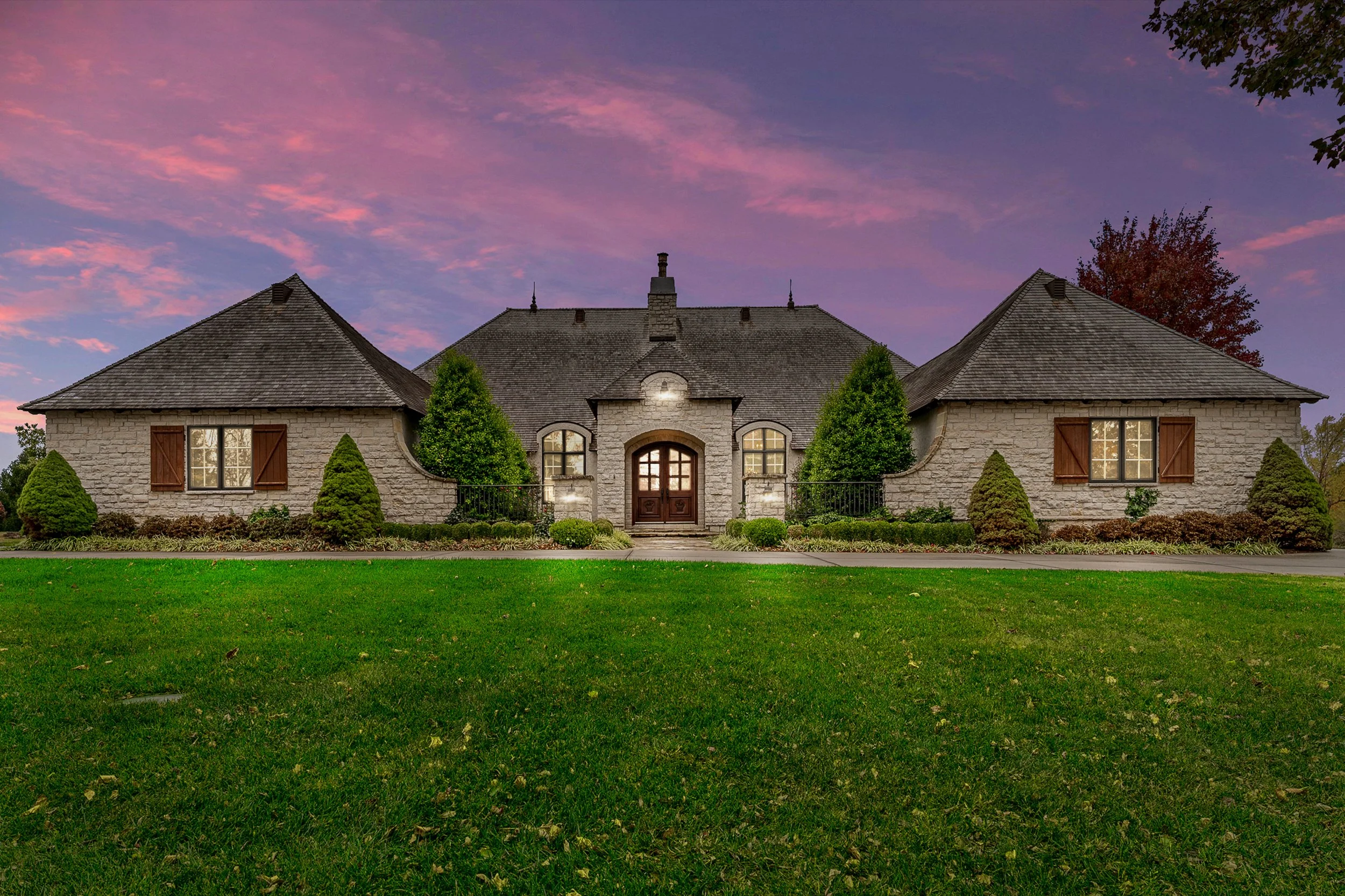A large stone house with a steeply pitched roof, surrounded by a well-maintained lawn and shrubbery, under a colorful sky at dusk.