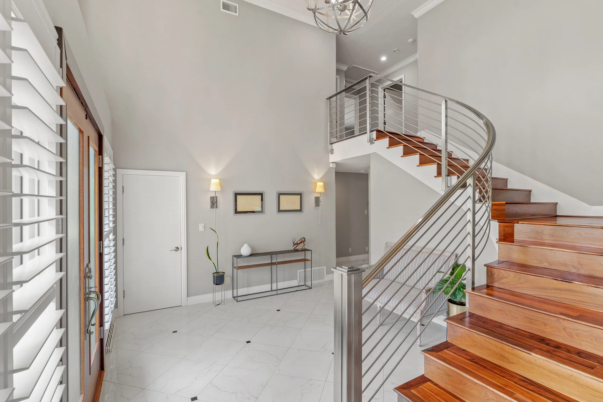 Interior view of a modern house entryway with a curved wooden staircase with metal railings, white walls, a marble floor with small black accents, a small table with decorative items, framed artwork, wall-mounted lamps, and a mixture of natural and a