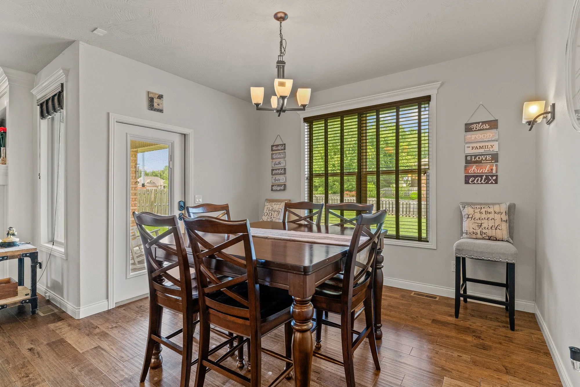 A dining room with a wooden table and chairs, large windows with blinds, a chandelier hanging from the ceiling, and decorative wall signs.
