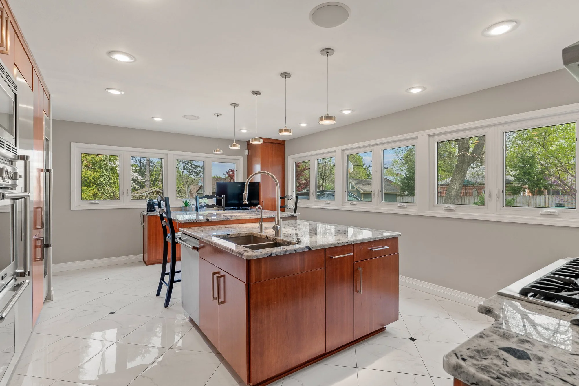 Modern kitchen with wooden cabinets, granite island, stainless steel appliances, white tiled floors, large windows showing green trees outside, and ceiling-mounted lights.