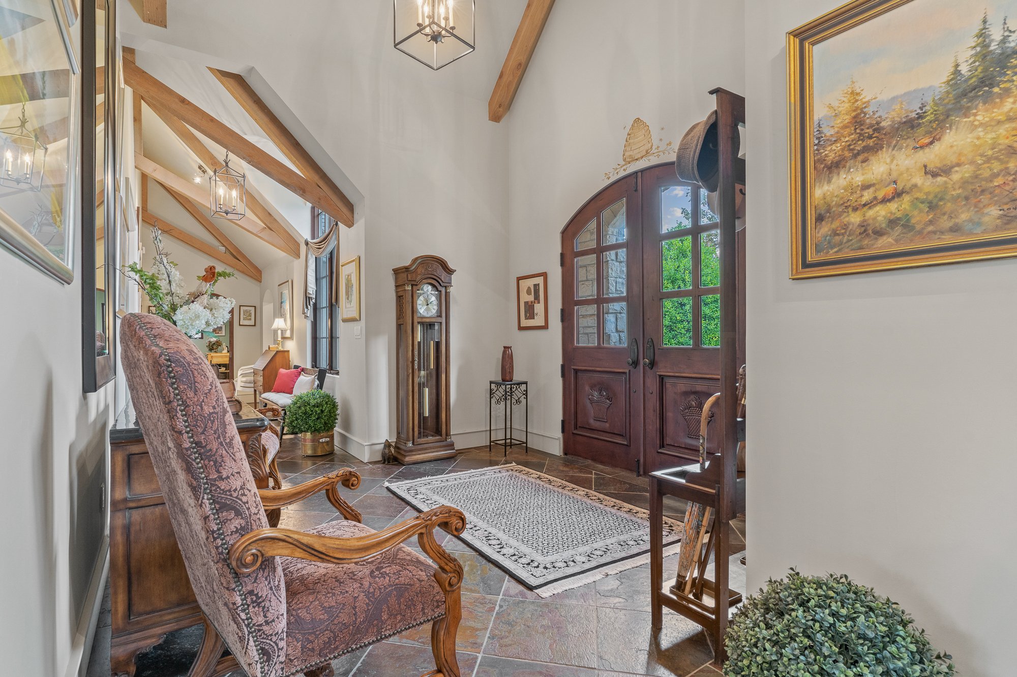 Entrance hall with wooden double doors, framed landscape painting, grandfather clock, floral arrangement, and vintage furniture.