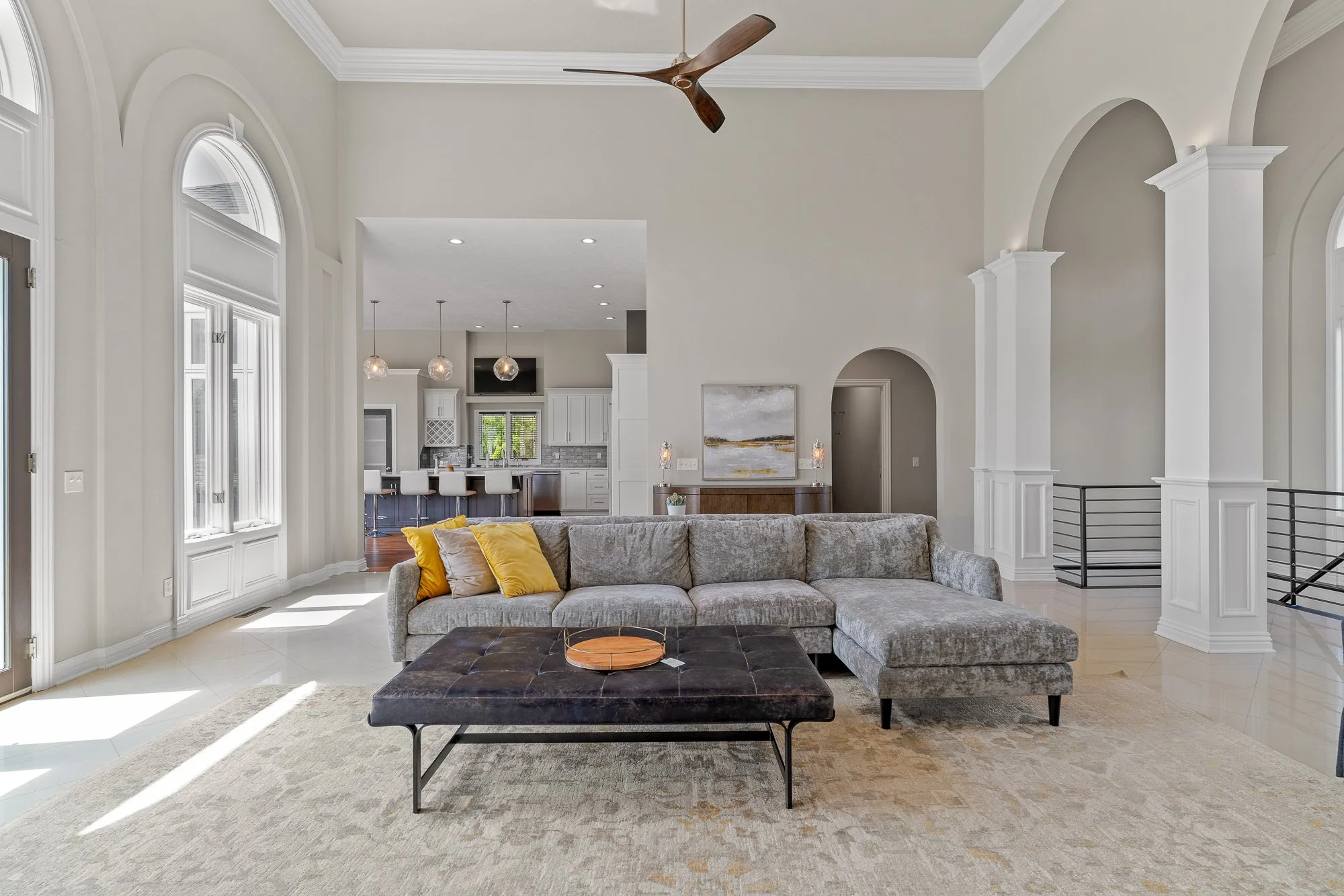 Living room with a large sectional sofa with yellow and beige pillows, a coffee table with a tray, high ceilings, arched windows, a ceiling fan, and an open kitchen in the background with pendant lights and a bar with white chairs.