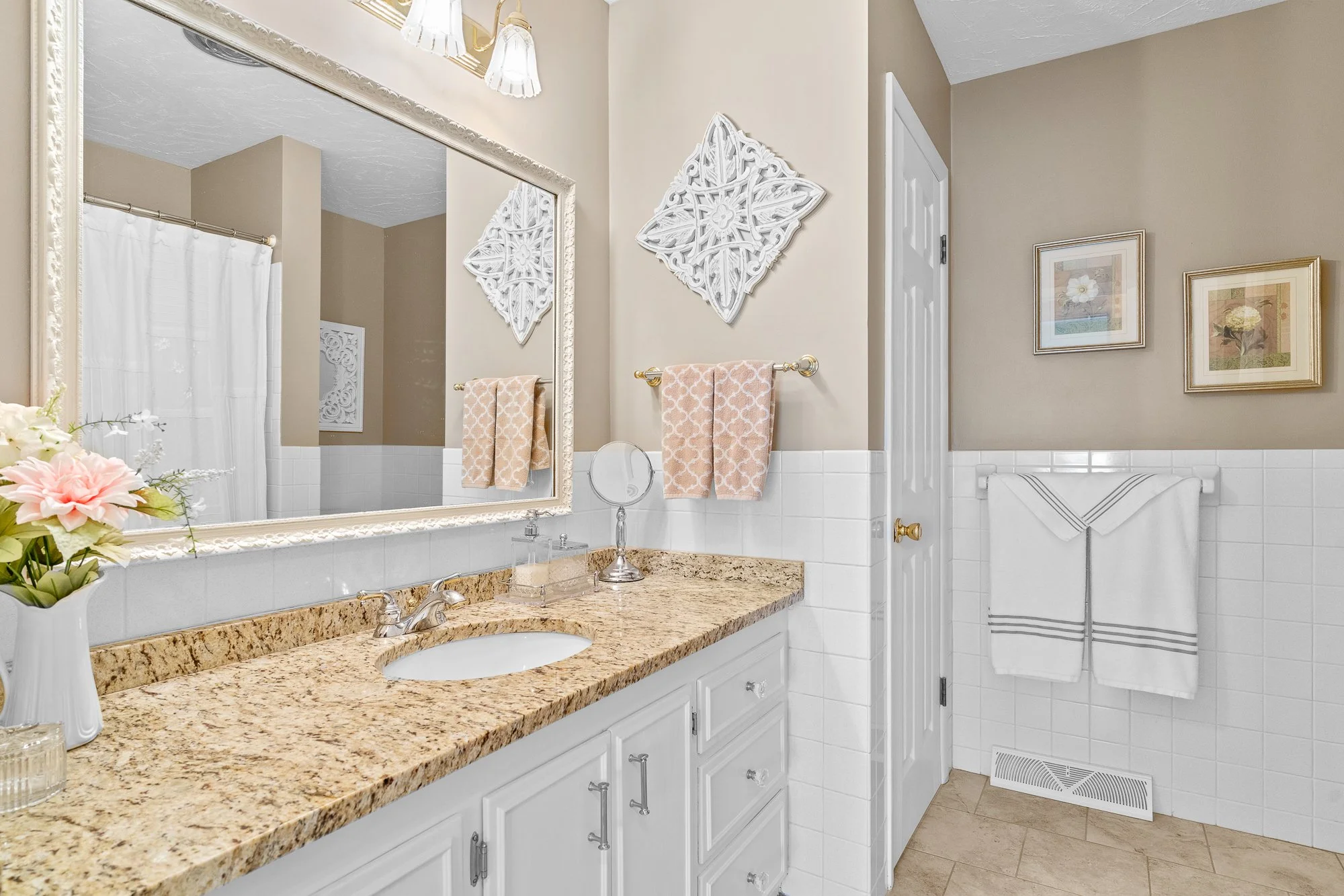 A bathroom vanity with a granite countertop, a faucet, and a large mirror. A vase with pink and white flowers is on the left side of the countertop. Two beige towels hang on a gold towel rack. The wall has framed floral artwork. There are decorative 
