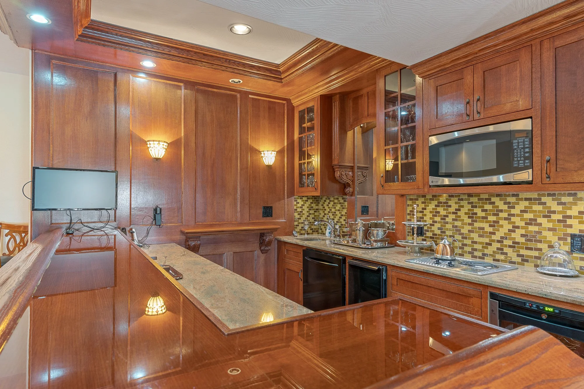 A kitchen with wooden cabinets, a granite countertop, and a mosaic tile backsplash. There is a microwave, a small television, and various kitchen appliances and utensils on the counter. Recessed ceiling lights and wall sconces illuminate the space.