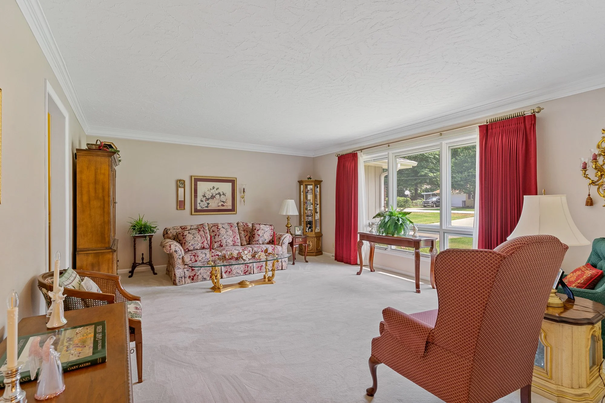 Living room with beige walls, white ceiling, large window with red curtains, floral sofa, armchair, wooden furniture, potted plant, and decorative items.