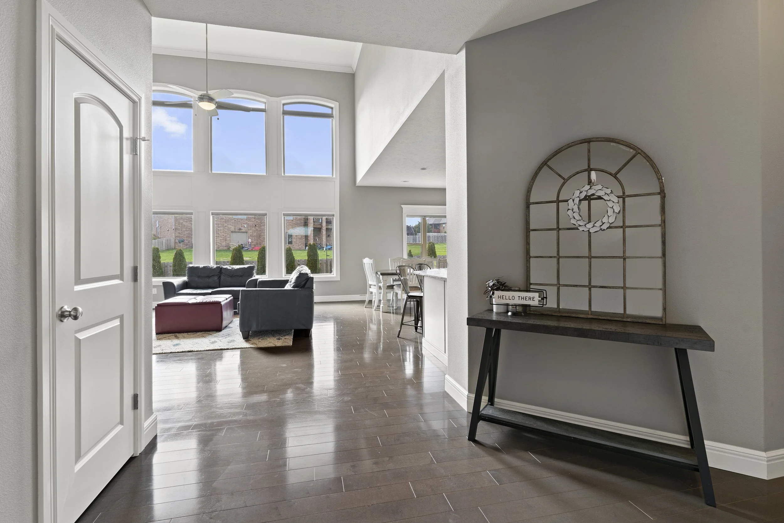 View from the entryway into a bright living room with large windows, a ceiling fan, a black sofa, a red ottoman, a dining table with chairs, and wall decor including a wreath and a sign that says "Hello There".