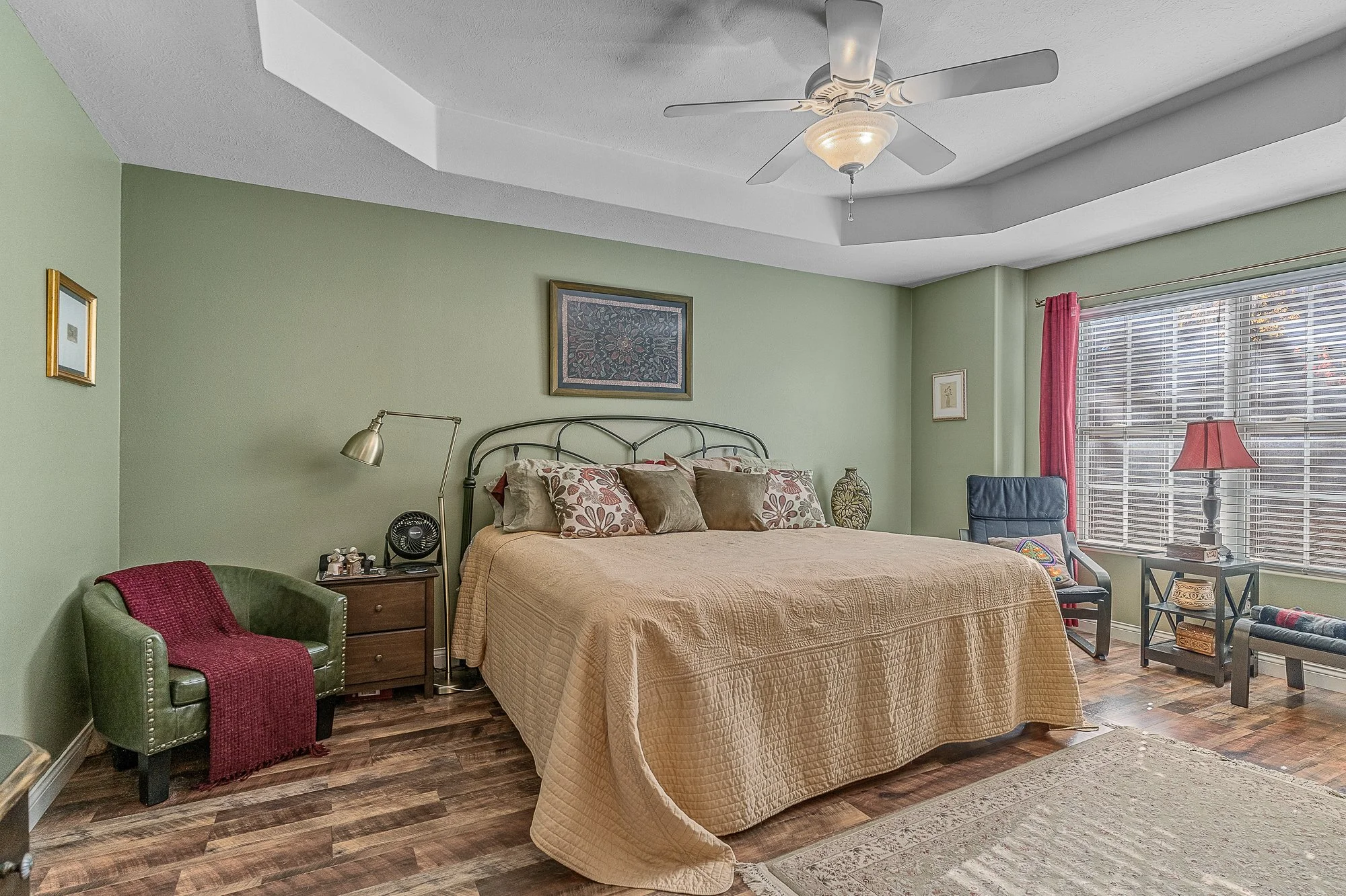 A bedroom with green walls, a beige bedspread, various decorative pillows, a black bedframe, side table with a small fan, a green armchair with a red blanket, a wooden nightstand, a tall lamp, a chair, a small table, and a large window with pink curt
