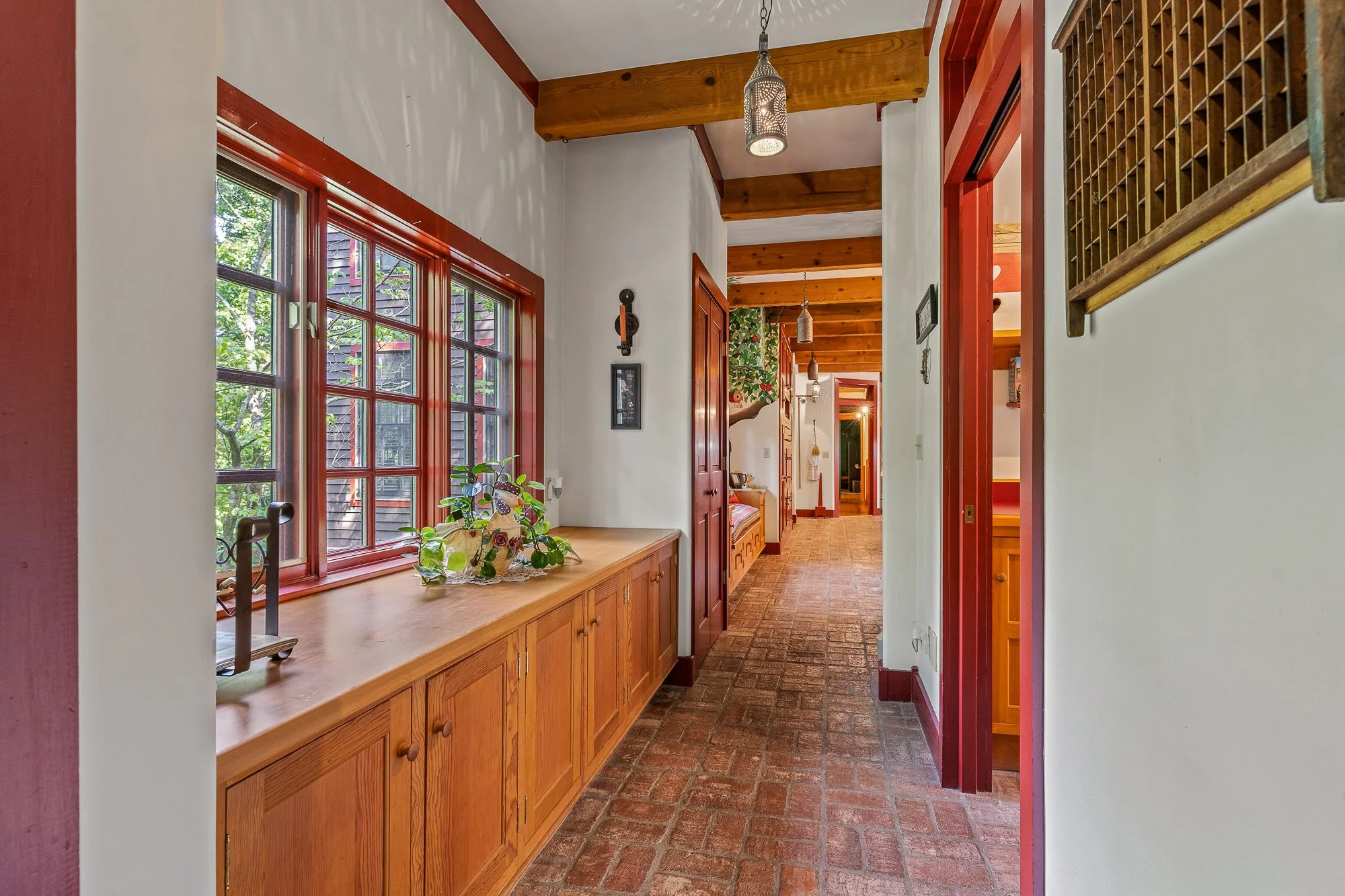 A hallway with white walls, wooden ceiling beams, brick flooring, red window frames, and a wooden built-in cabinet with a plant on top. Multiple pendant lights hang from the ceiling, and there is artwork and decor on the walls.