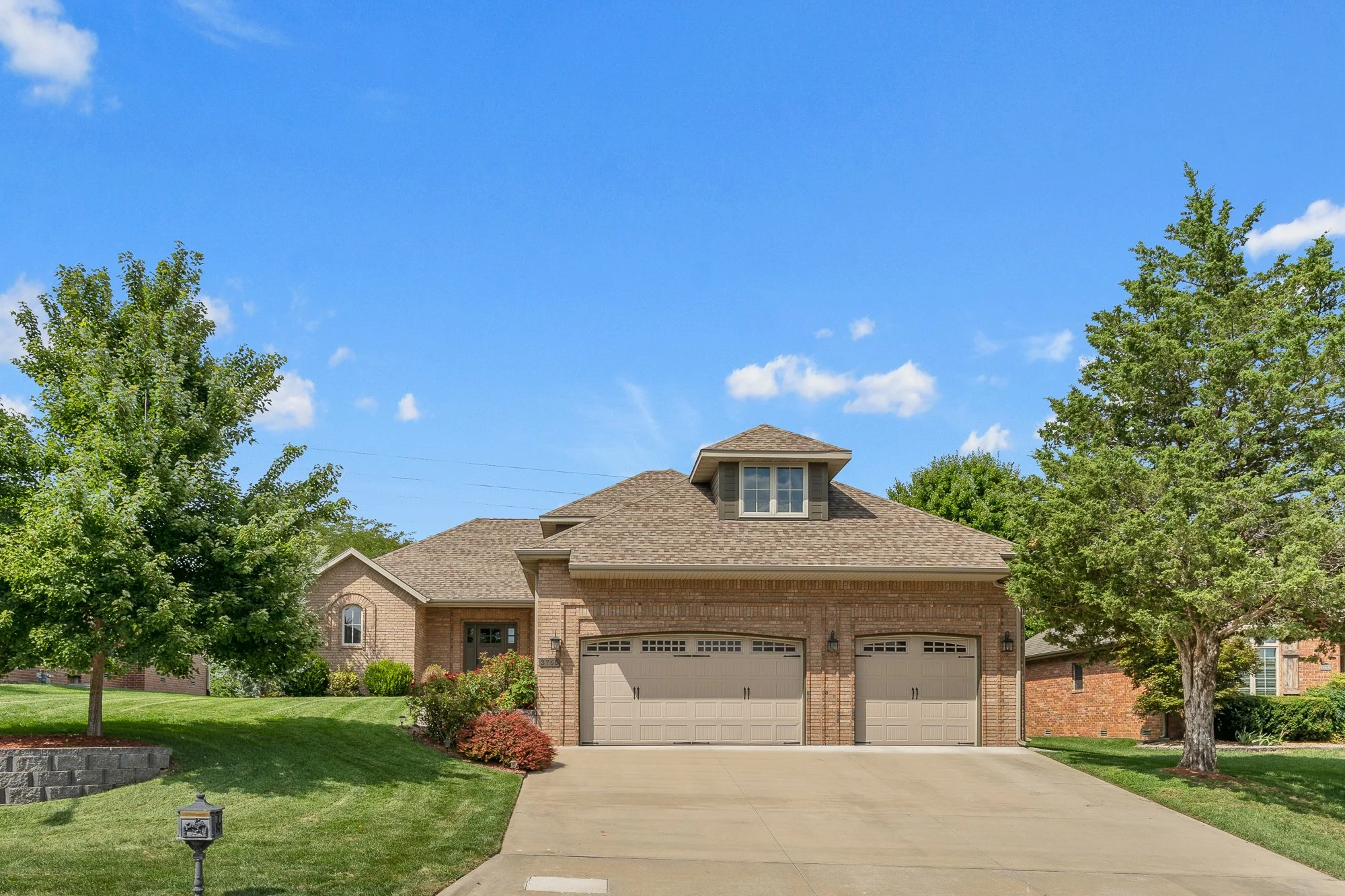 A brick house with a two-car garage, a driveway, and two trees in the yard. There are shrubs and flowers near the house, and a small lamp post on the lawn. The sky is blue with a few clouds.