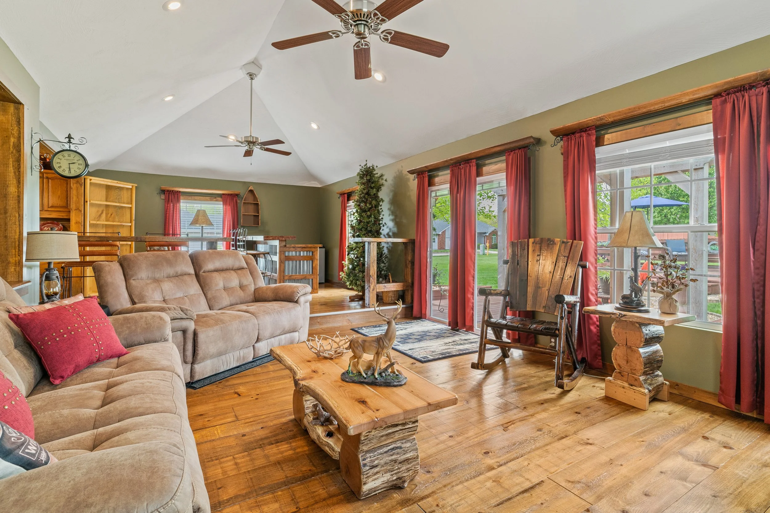 Living room with a beige sectional sofa, wooden coffee table with a deer figurine, large windows with red curtains, ceiling fans, and hardwood floors.
