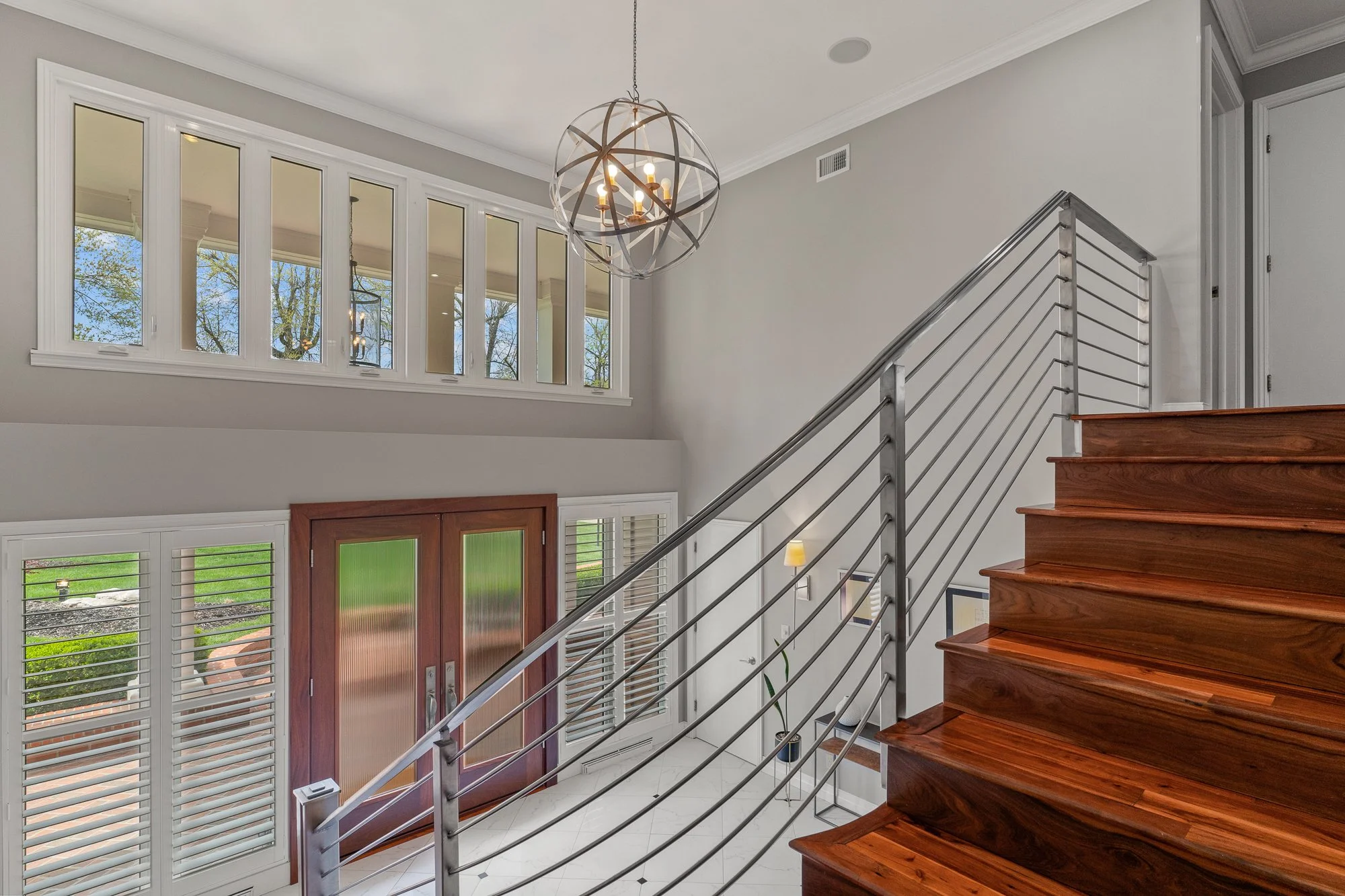 Entryway of a house with a staircase, wooden steps, metal railing, and a glass front door. Large windows with white shutters let in natural light, and there is a spherical chandelier hanging from the ceiling.
