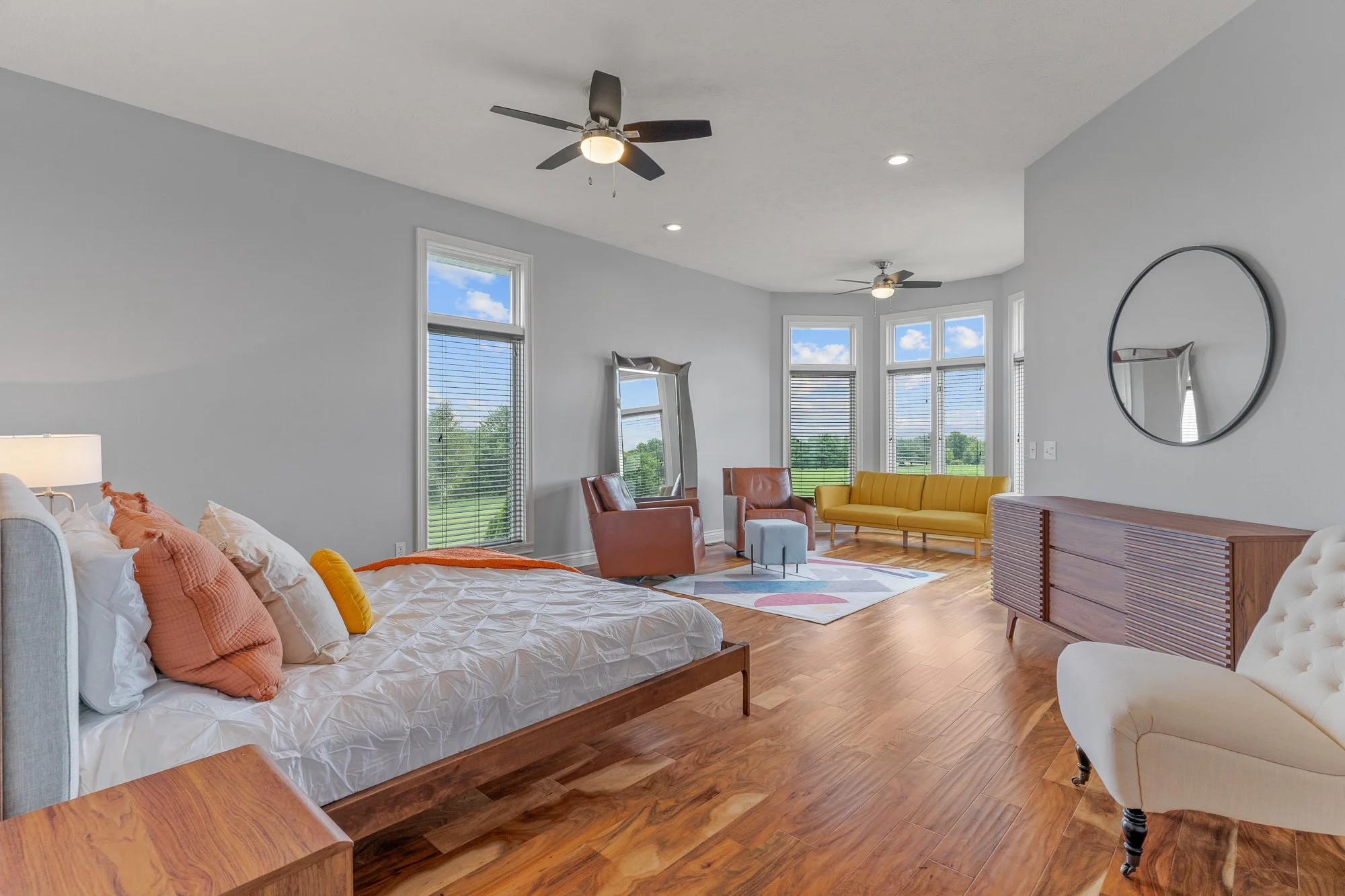 Living room with large windows, wooden floor, colorful furniture, and modern decor, including a bed, chairs, a yellow sofa, and wall mirror.