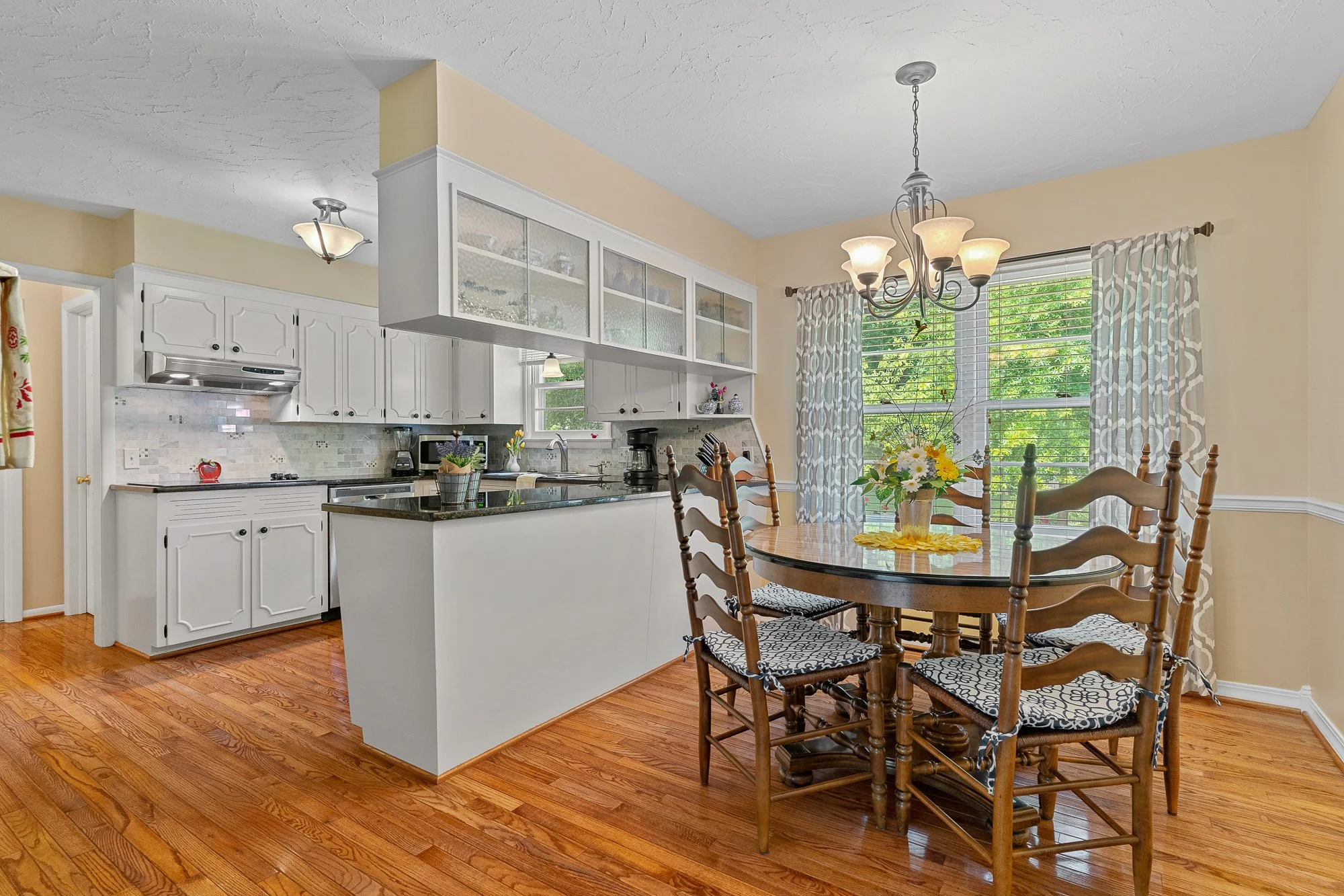 A kitchen and dining area with white cabinets, a black countertop, a wooden dining table with six chairs, a vase with yellow and white flowers, large windows with patterned curtains, hardwood floors, and a ceiling light fixture.
