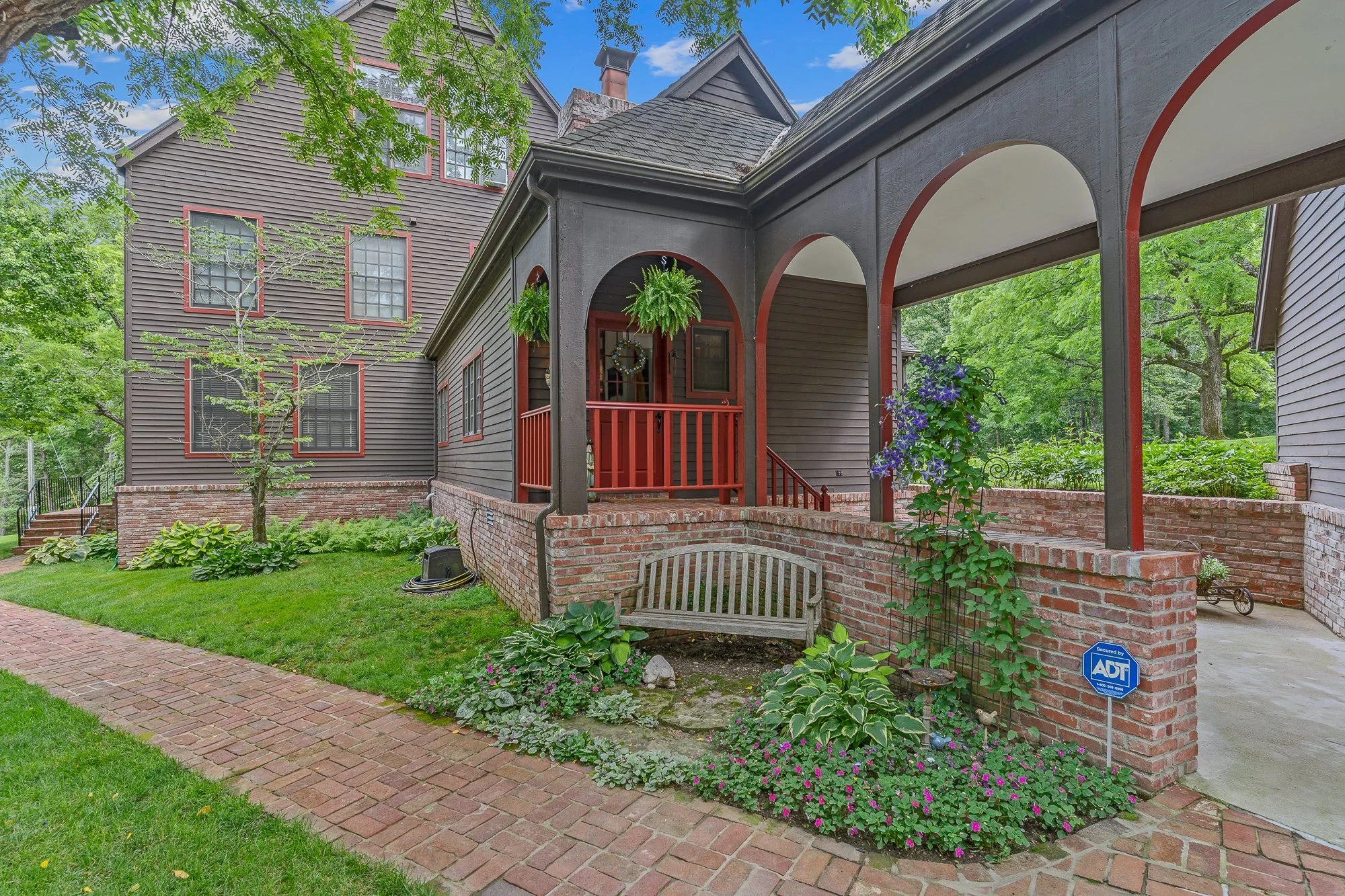The image shows the front porch of a house with black wood siding, red trim around windows and the porch door, a brick foundation, a small set of stairs leading to the entrance, decorated with hanging plants. There’s a garden with plants and flowers,