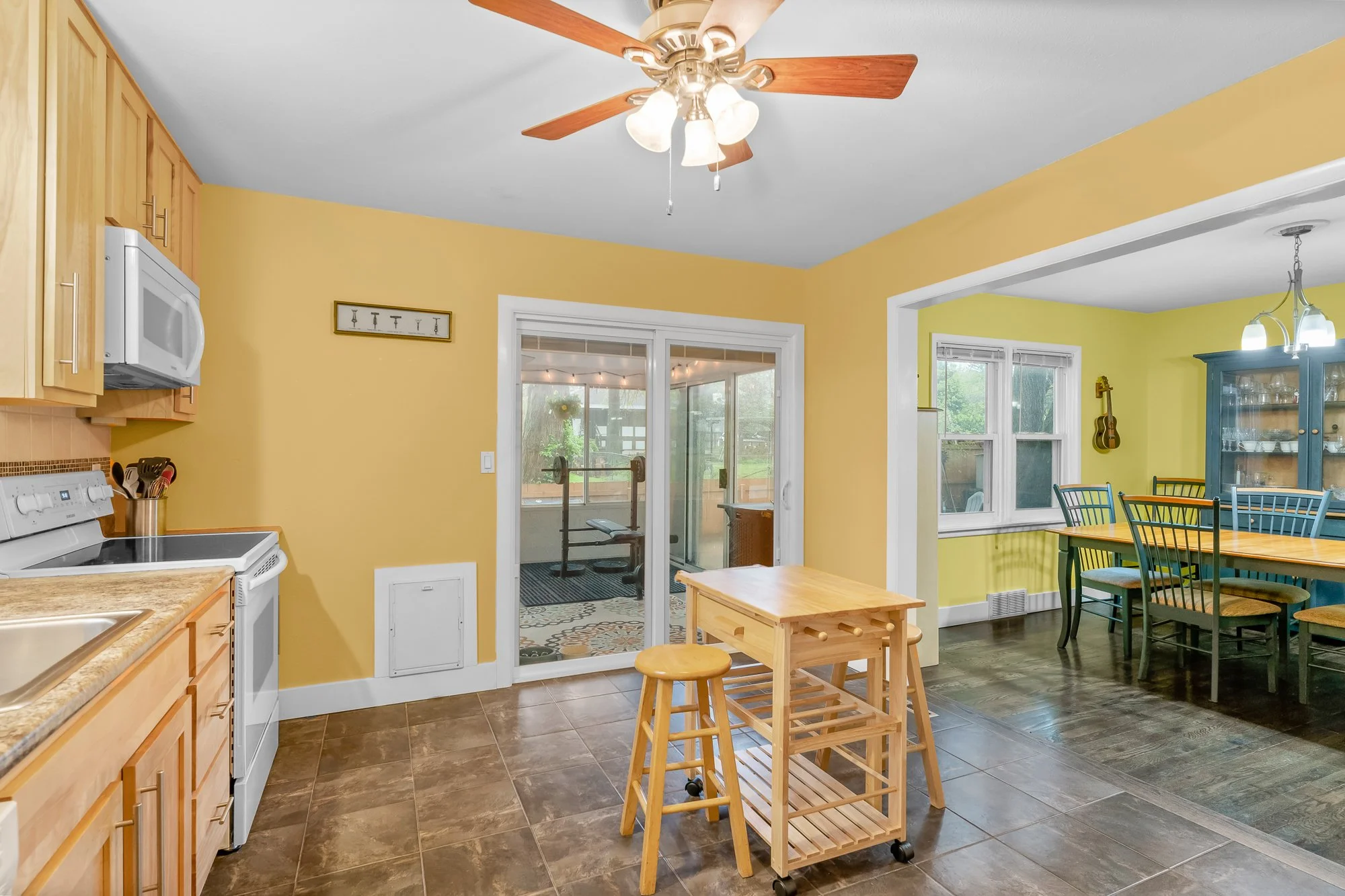 Kitchen with yellow walls, wooden cabinets, a white stove, a microwave, and a small wooden cart with stools. There is a doorway leading to a sunroom or porch with a weightlifting bench. A dining area with a green and yellow color scheme and a guitar 