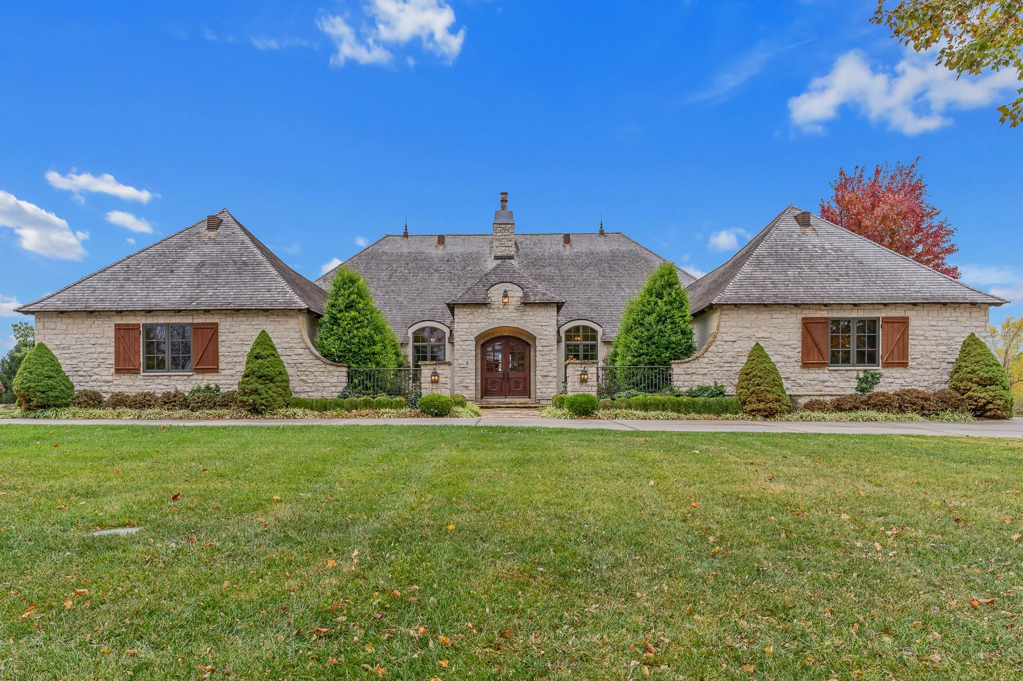 A large stone house with a peaked roof, dark wooden shutter windows, and a lush front yard with trimmed bushes and a green lawn.