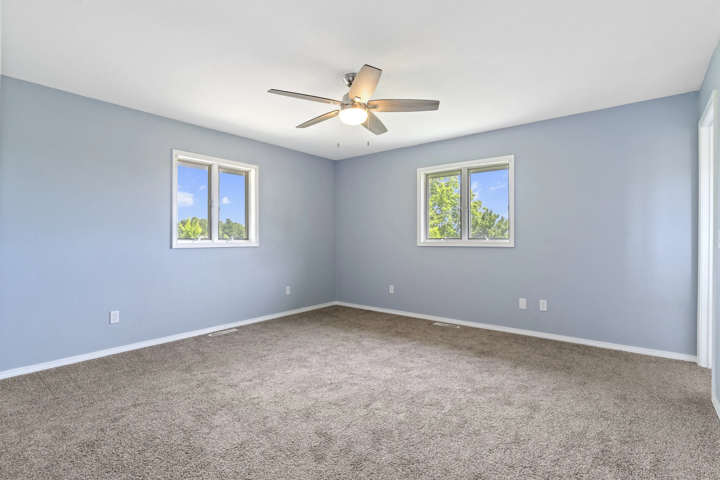 Empty room with light blue walls, two windows with views of green trees and blue sky, ceiling fan with light, brown carpet, and white baseboards.