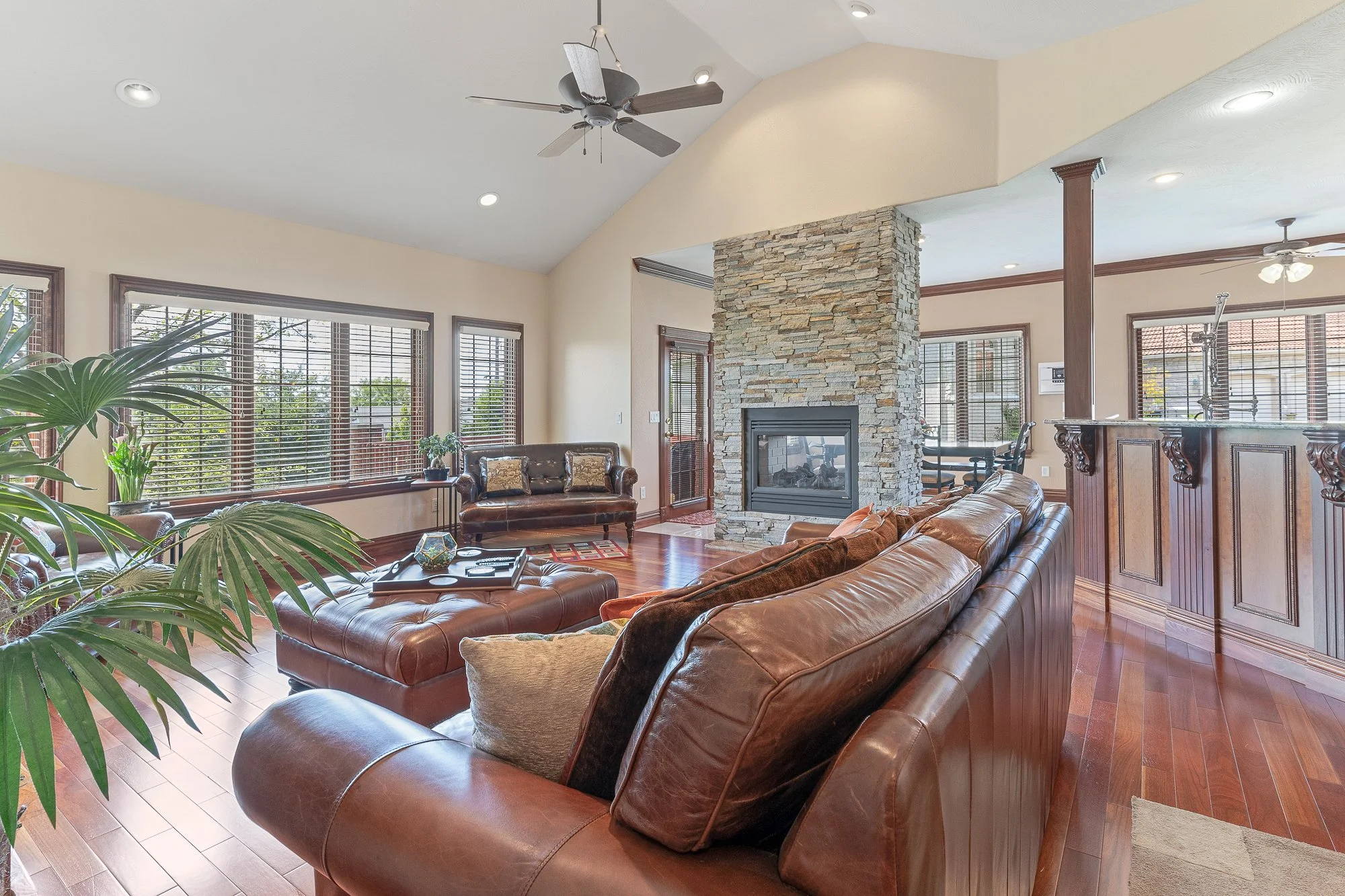 Spacious living room with large windows, leather sofas, a stone fireplace, ceiling fans, and wooden accents.