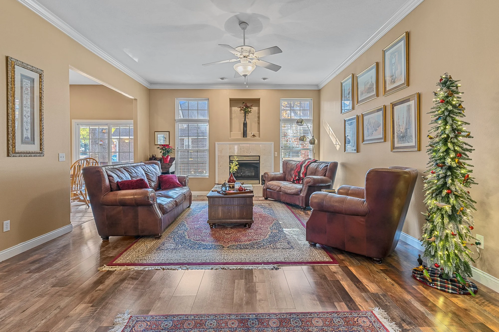 Living room with beige walls, hardwood floor, leather sofas, a fireplace, framed pictures on the wall, a Christmas tree, and windows letting in natural light.