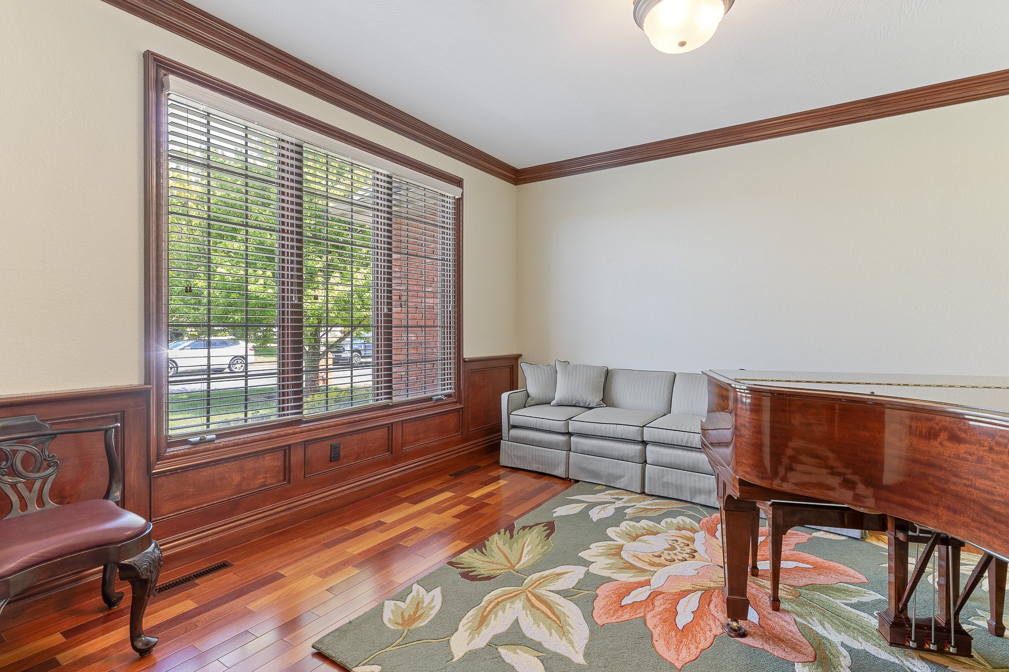 Living room with large window, cream walls, wooden crown molding, hardwood floor, gray sofa with throw pillow, area rug with floral pattern, grand piano, and a chandelier ceiling light.