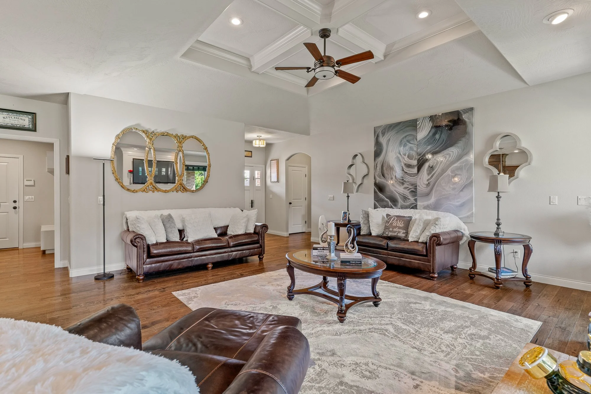 Living room with white walls, vaulted ceiling with a fan, two dark brown leather sofas with white and gray pillows, oval wooden coffee table, decorative mirrors, abstract artwork, lamps, and hardwood flooring.