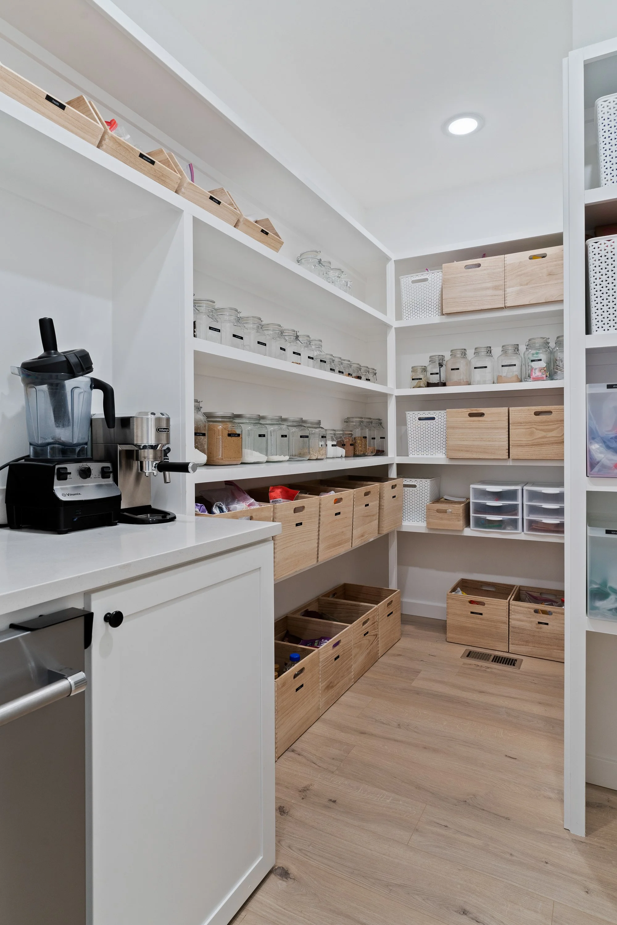 A pantry with white shelves containing glass jars, wooden boxes, and white baskets. A countertop with a blender, coffee maker, and small storage drawers. Wooden flooring and an air vent on the floor.