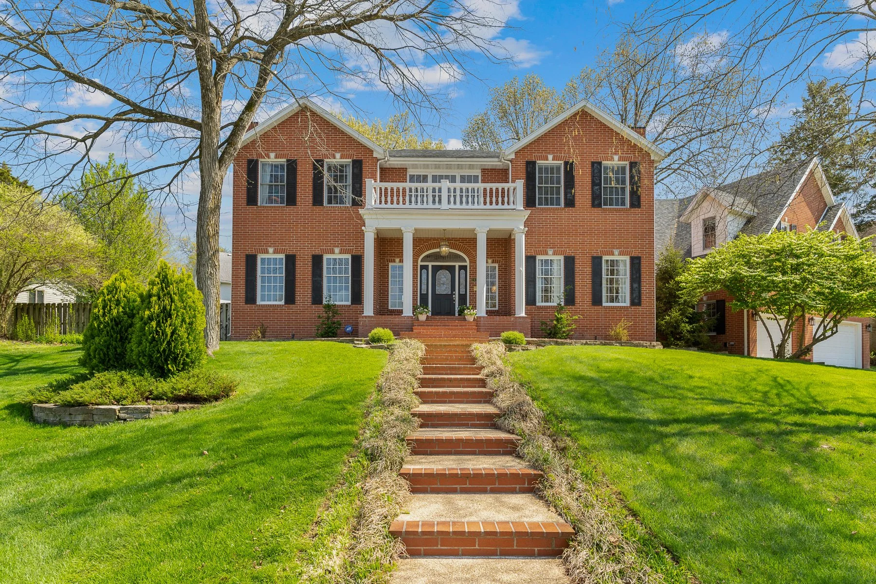 A large brick house with a front porch, black shutters, and a balcony, surrounded by a well-maintained green lawn and landscaping, with trees and a blue sky in the background.