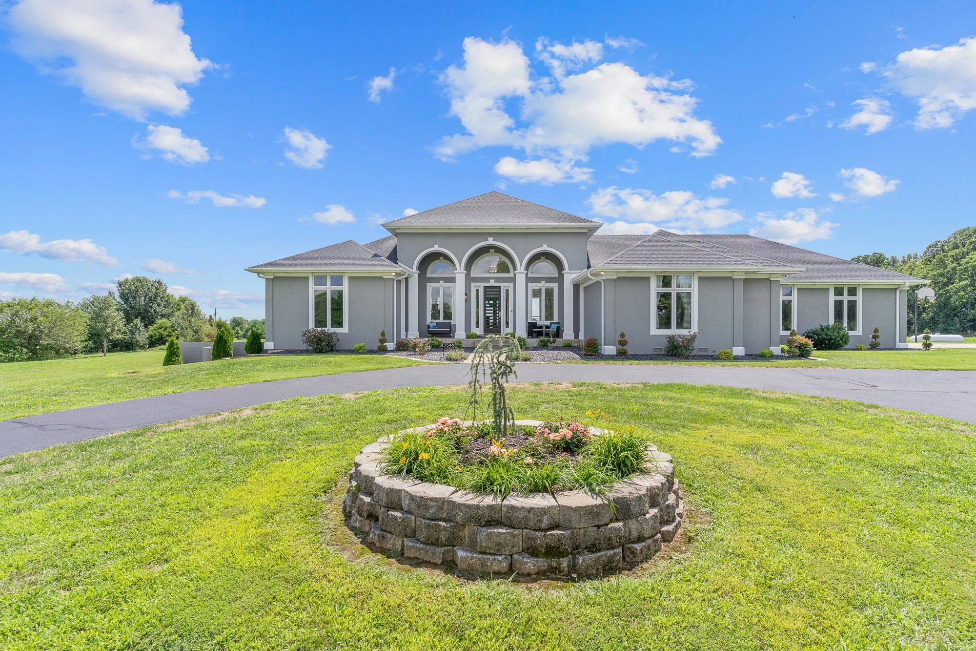 Large gray house with a well-maintained lawn, flower bed with pink and yellow flowers, and a circular flower bed in the foreground. The house has multiple arched windows, a front porch with seating, and a gabled roof. The sky is blue with scattered c