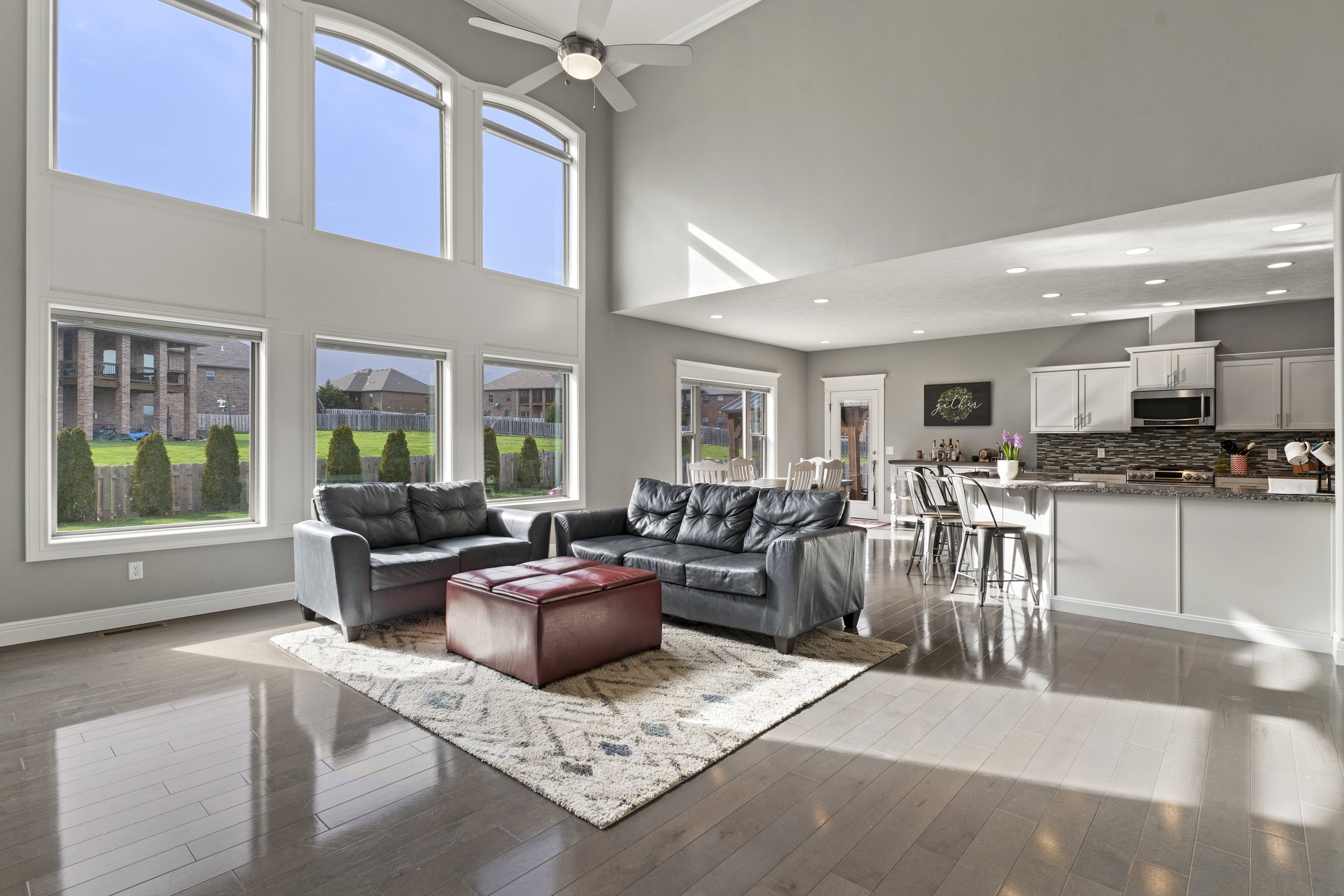 Living room with large windows, gray walls, dark hardwood floors, black leather couches, a red ottoman, and a view of neighboring houses and a backyard with green grass and shrubs.