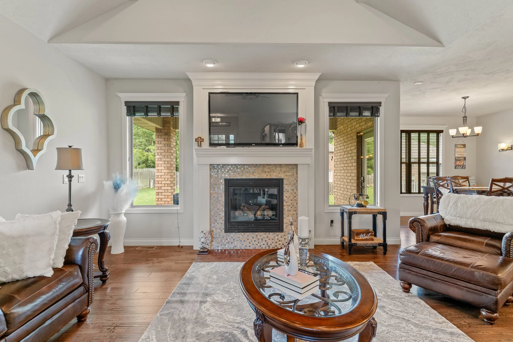 Living room with a decorated fireplace, flat-screen TV above, brown leather sofas, a glass coffee table, large windows, and a dining area in the background.