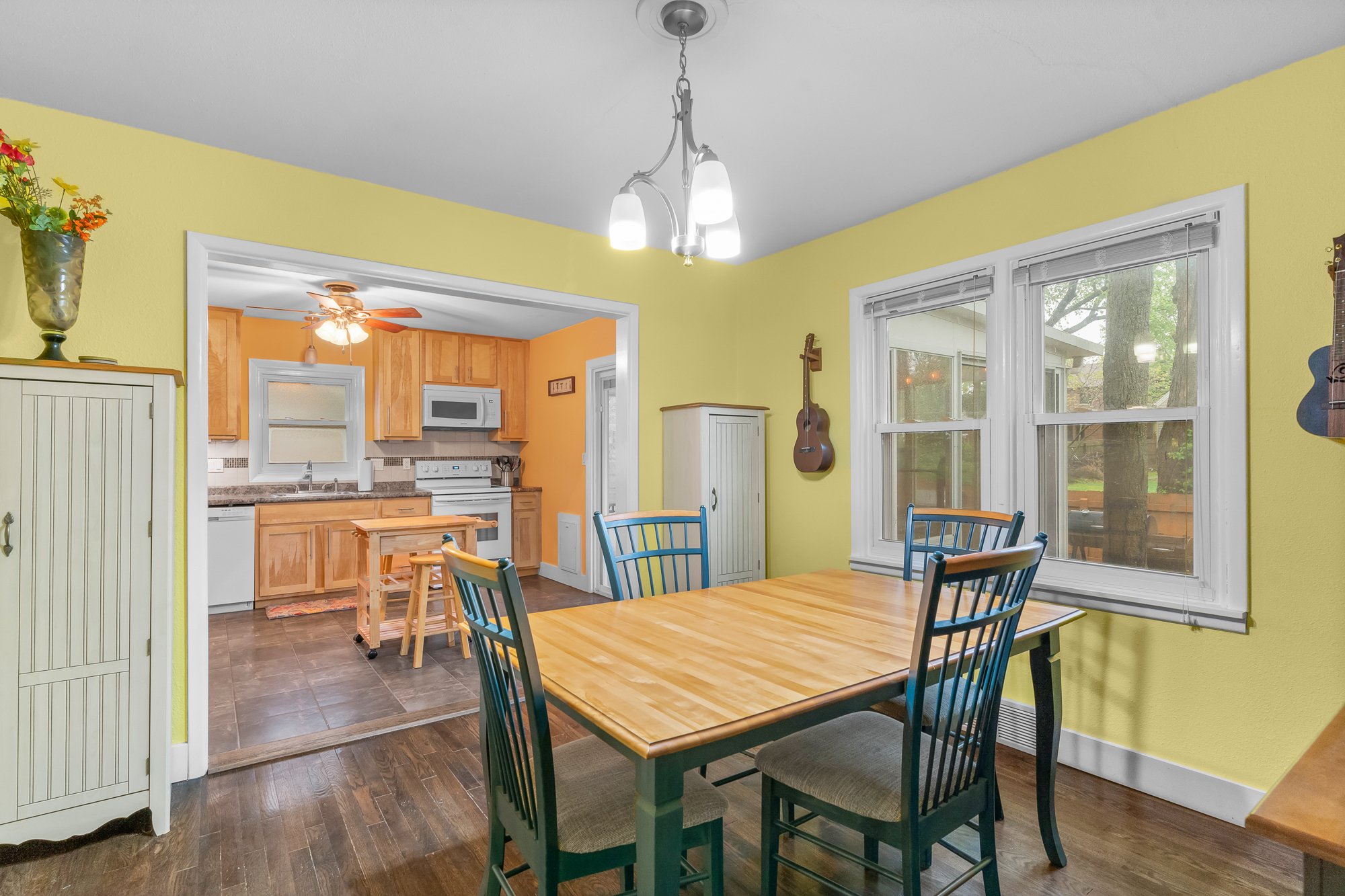 Interior of a bright kitchen and dining area with a wooden table and chairs. The kitchen has wooden cabinets, a stove, and a microwave. A large window shows outdoor greenery. Decorations include a guitar on the wall and a vase of flowers.