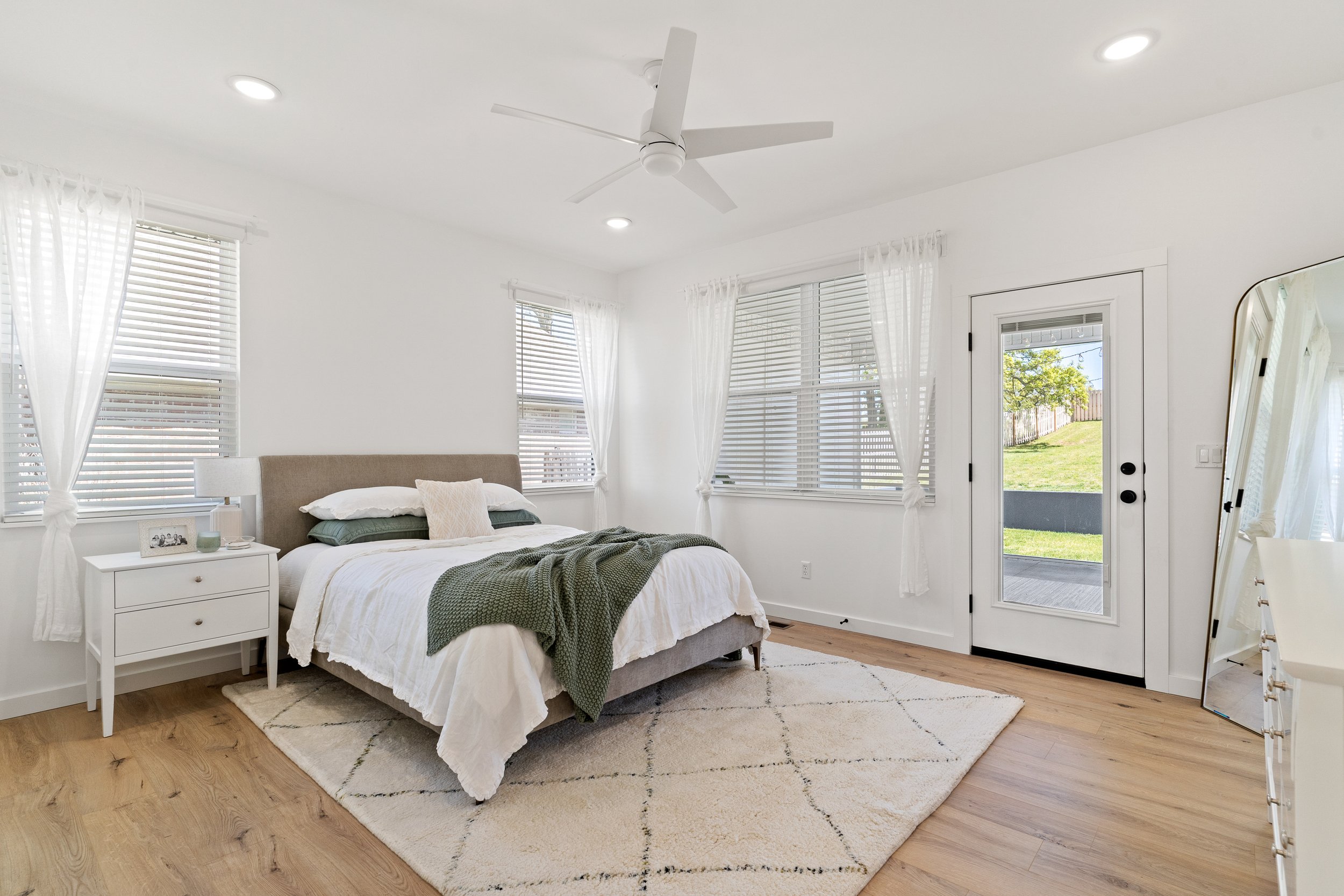 Bright, cozy bedroom with white walls, large windows with sheer curtains, a large bed with white linens and green accents, a white nightstand with a photo frame and lamp, a mirror, and a door leading outside to a grassy yard.