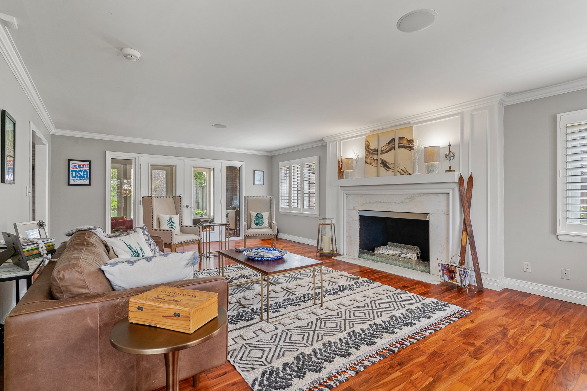 A living room with a brown leather sofa, beige armchairs with green leaf pillows, a wooden coffee table, a fireplace with decorative items on mantel, hardwood floor, and windows with shutters.