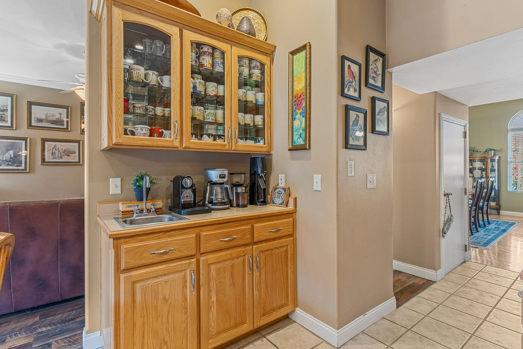 Kitchen corner with wooden cabinet with glass doors filled with mugs, a coffee maker, and a small countertop with a sink and a small framed picture. To the right, there's a wall with framed bird artwork, and an adjacent dining room with a rug, chairs