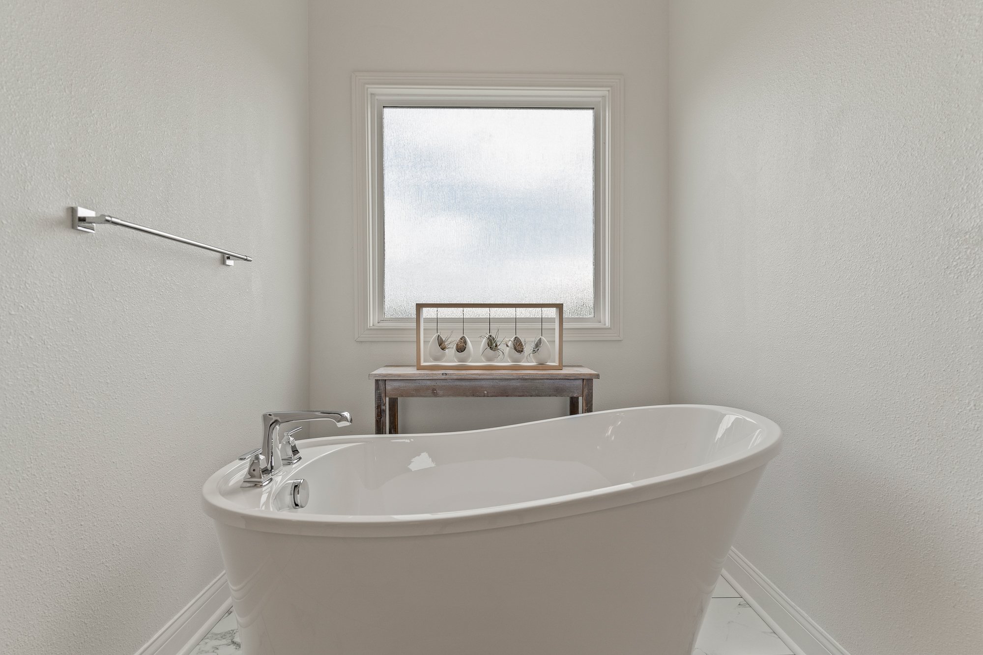 A white freestanding bathtub with silver fixtures in a bathroom with white walls. There is a large frosted window above the tub, and a wooden table holding small potted plants. A towel bar is mounted on the wall to the left of the window.