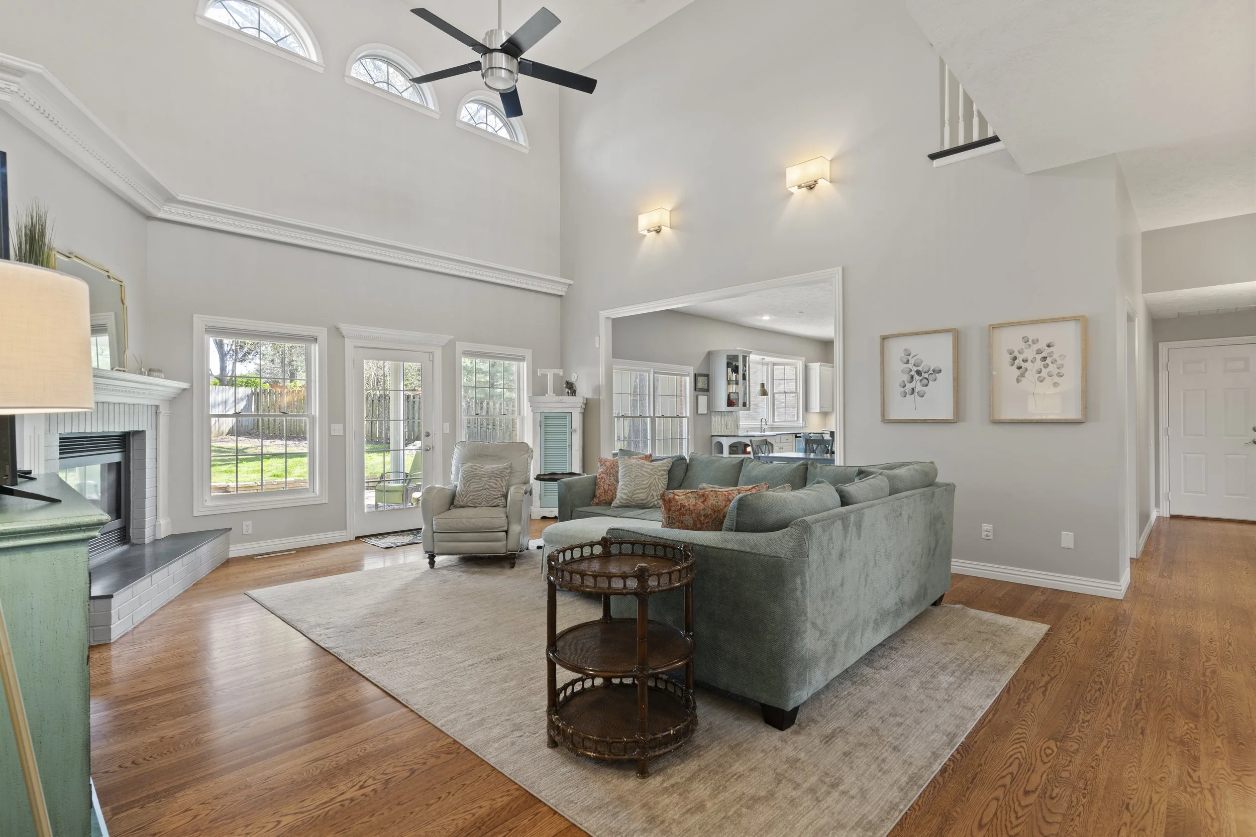 A spacious living room with high vaulted ceilings, large windows, and a ceiling fan. The room features a fireplace with a mantle, a green sofa with decorative pillows, an armchair, and a round wooden side table. The living area opens to a kitchen wit