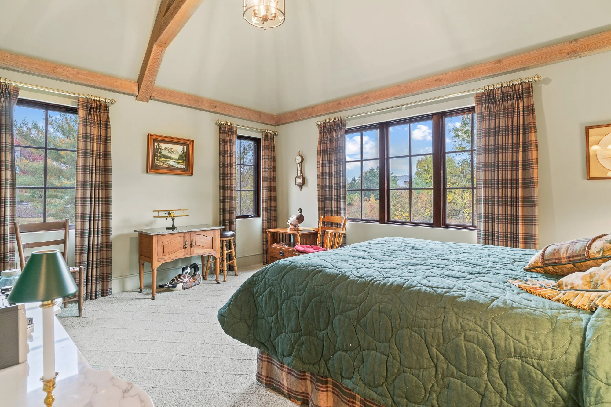 Bright bedroom with large windows, plaid curtains, green quilt on bed, vintage wooden furniture, picture on wall, and decorative duck sculpture.
