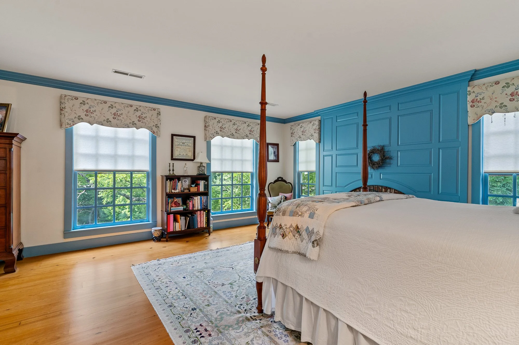 A bedroom with a four-poster bed, a blue panelled wall, three windows with floral valances, a bookshelf, framed pictures, and a cream-colored area rug.