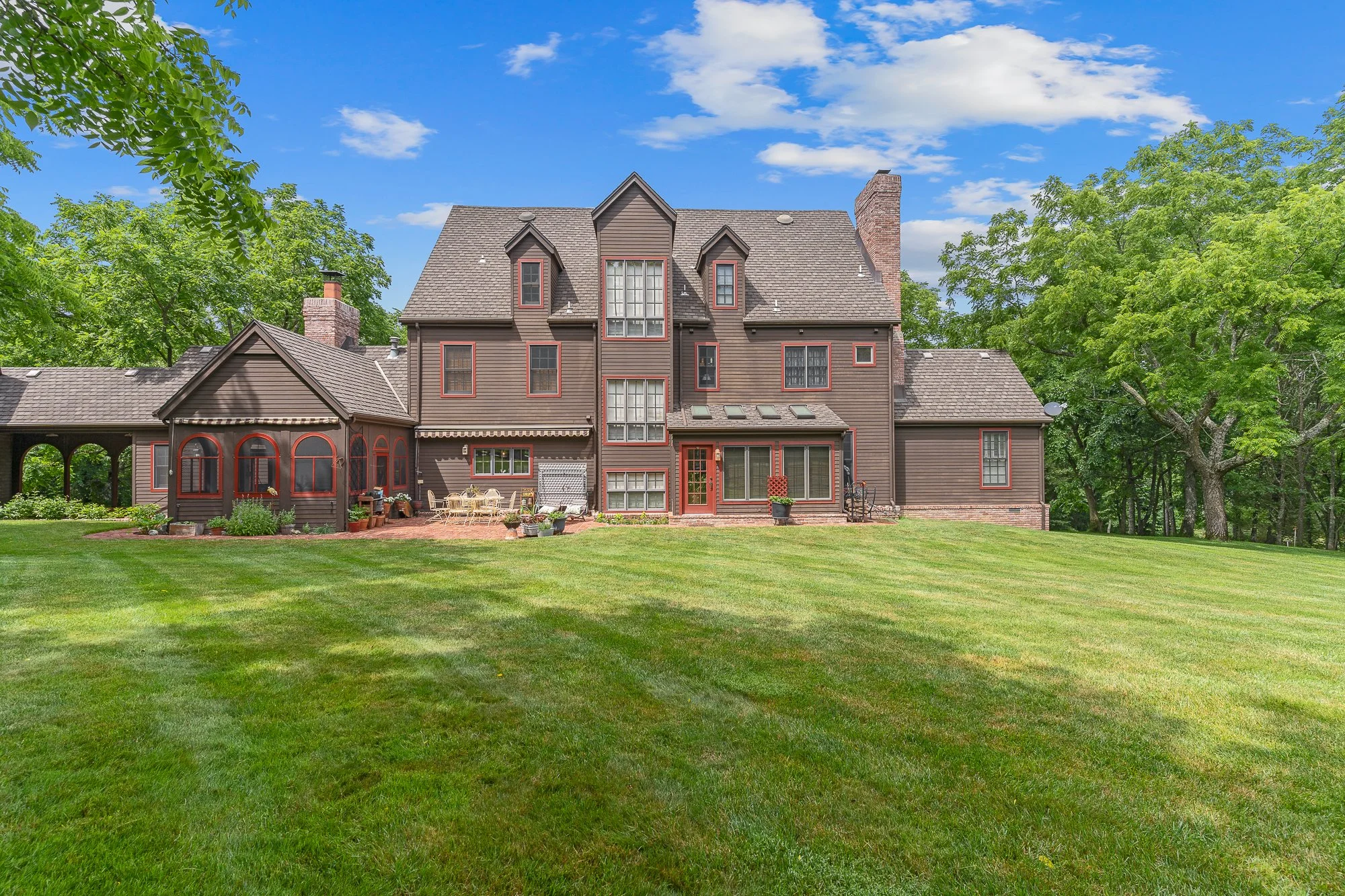Brown multi-story house with a gabled roof, brick accents, and large windows, surrounded by green lawn and trees, under a partly cloudy blue sky.