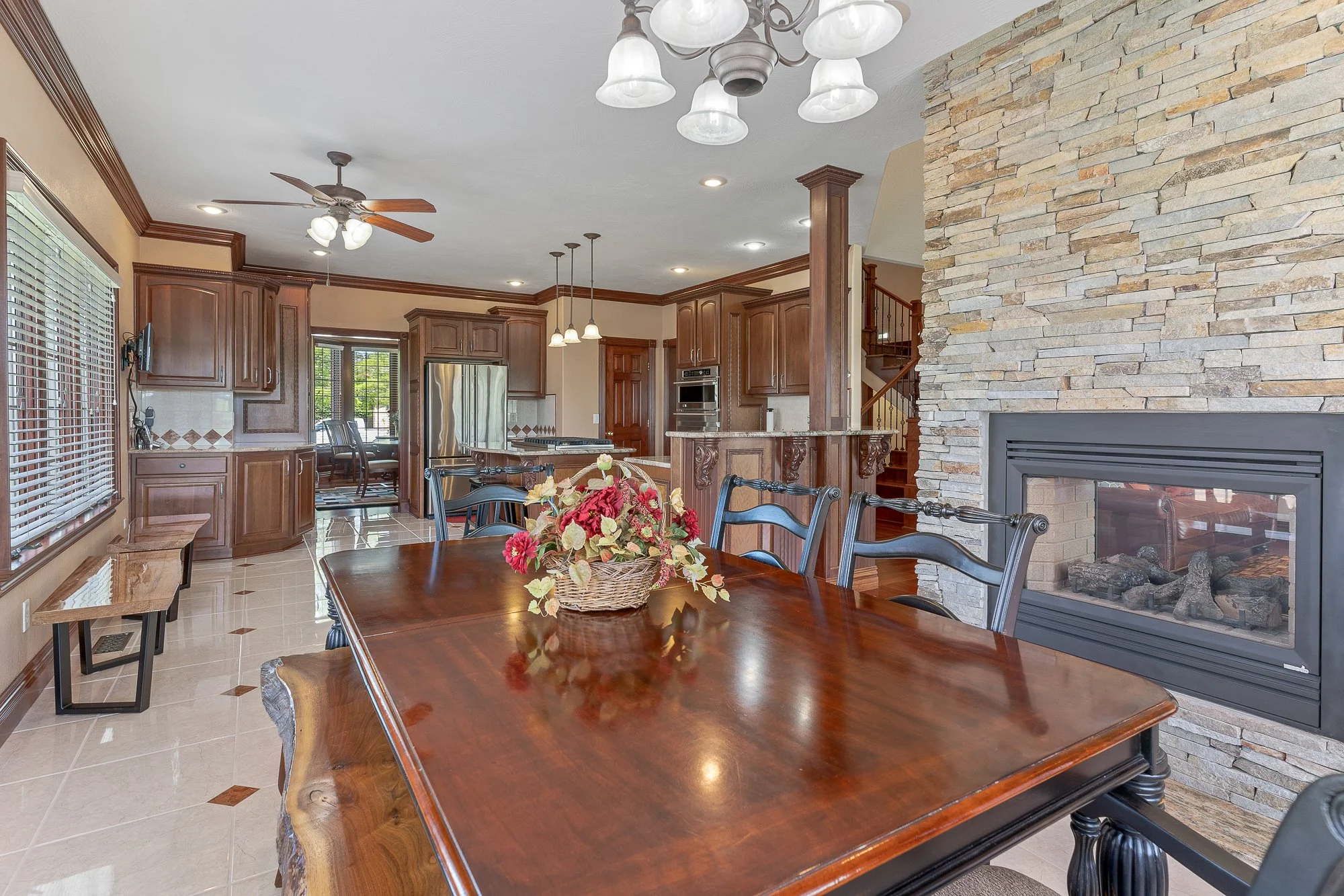 A dining room with a large wooden table, black chairs, a flower arrangement, a stone fireplace, wooden kitchen cabinets, a ceiling fan, and pendant lights in an open floor plan.