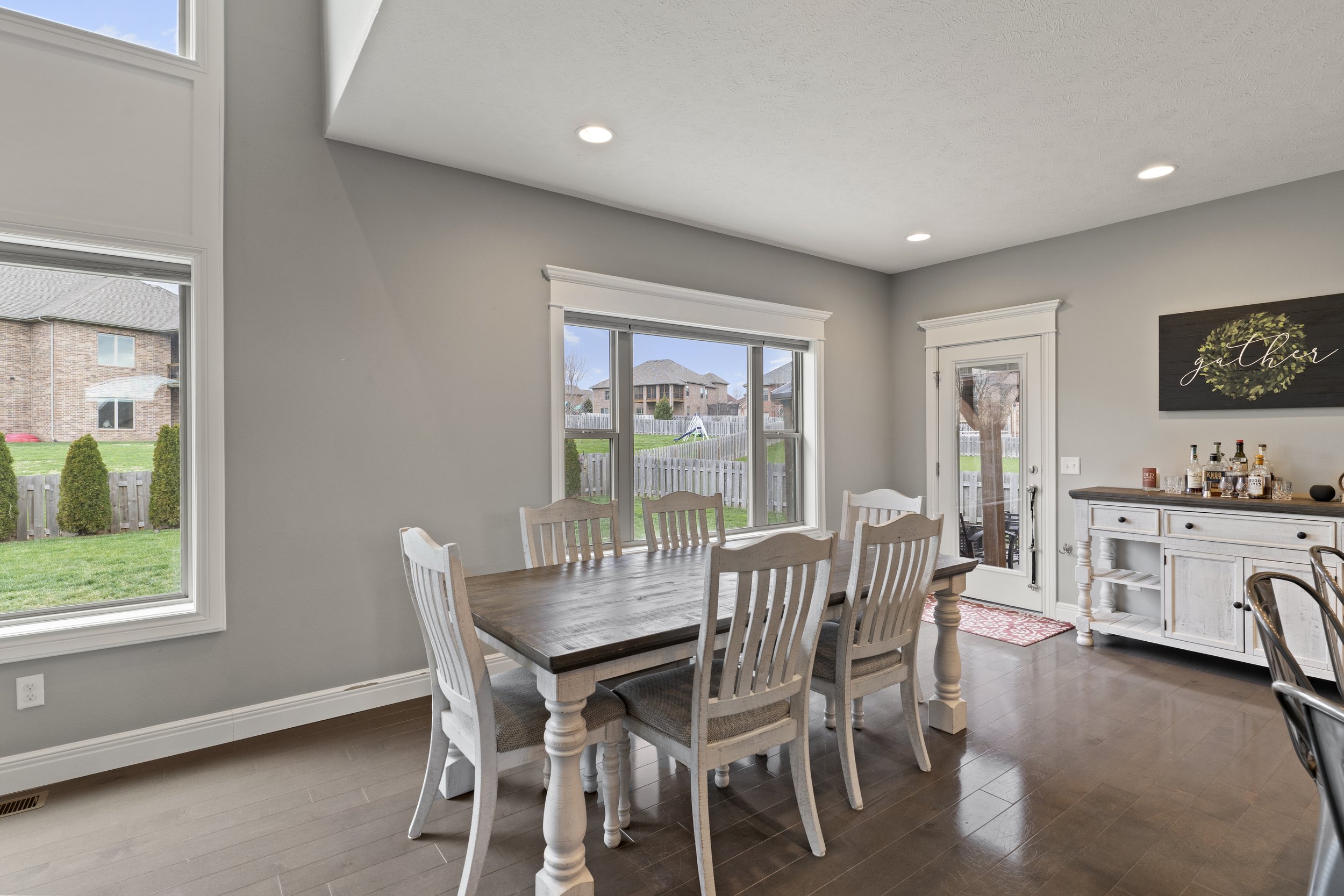 Living room with large windows, a wooden dining table with chairs, a white cabinet with bottles on top, and wall art with a wreath design.