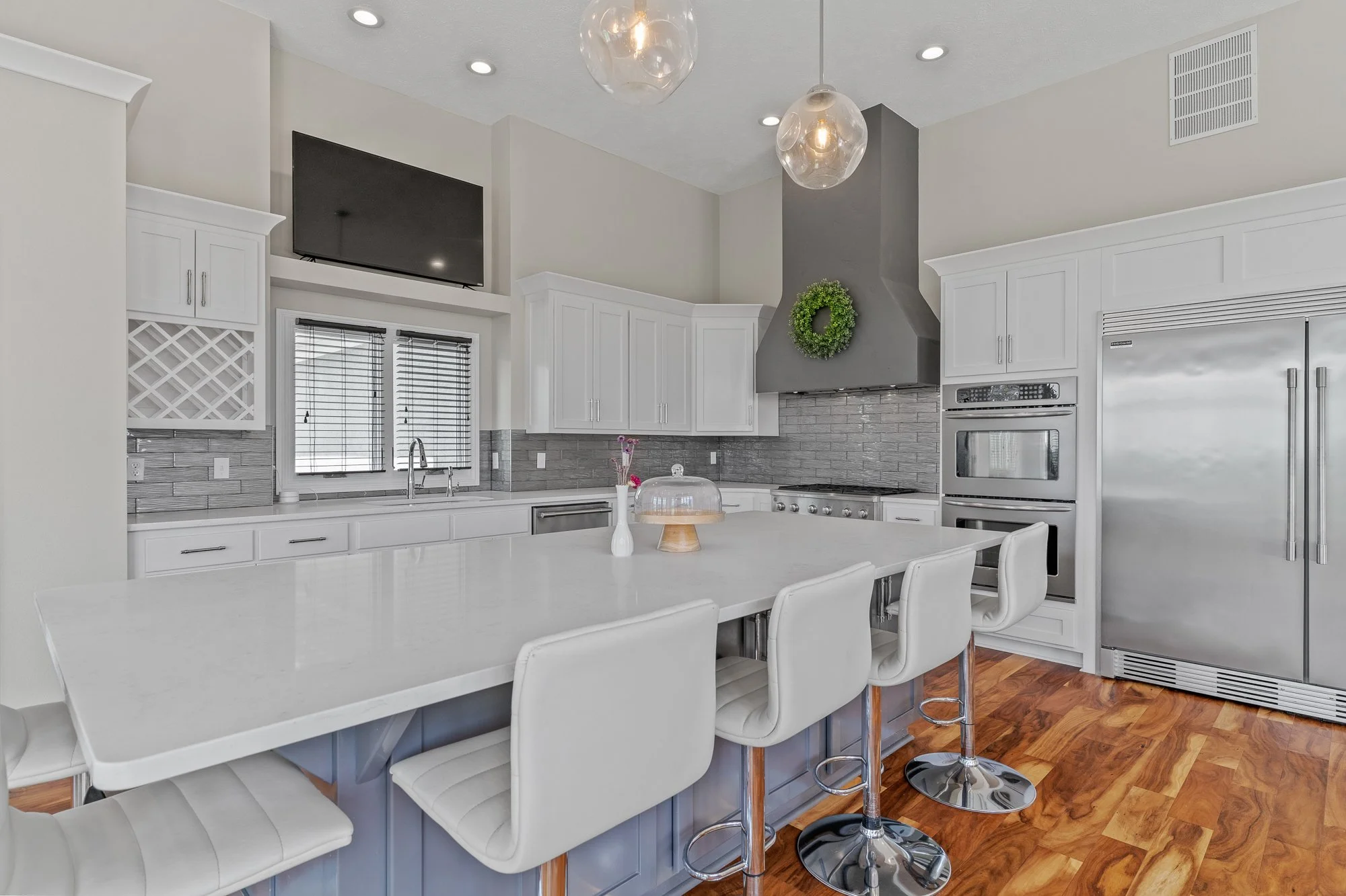 Bright modern kitchen with white cabinets, stainless steel appliances, a large island with white chairs, pendant lights, and a flat-screen TV mounted above the window.