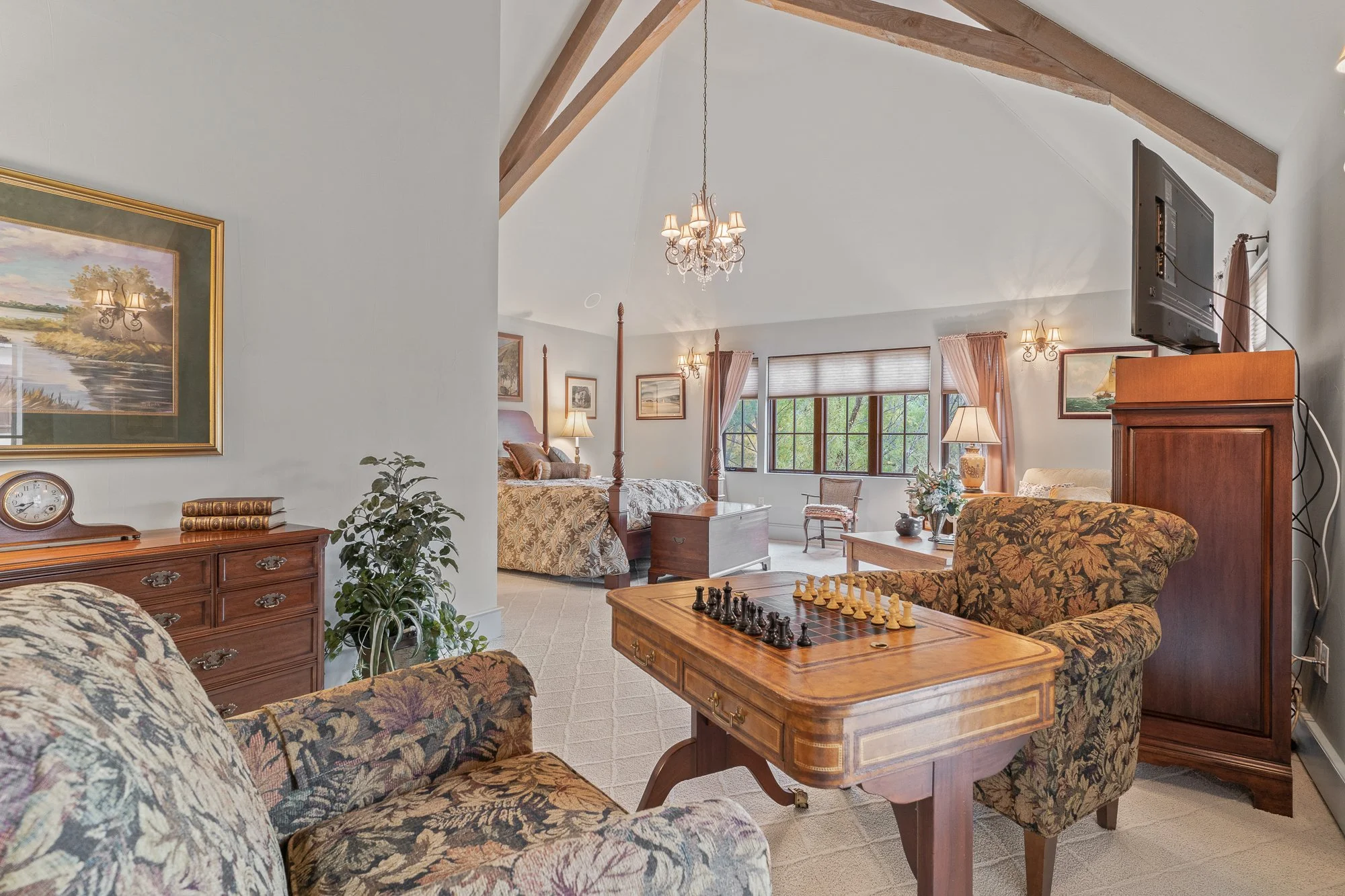 A cozy living room and bedroom area with floral upholstered furniture, a wooden chest with a chessboard, bedside lamps, framed landscape paintings, a crystal chandelier, and a large window with curtains showing trees outside.