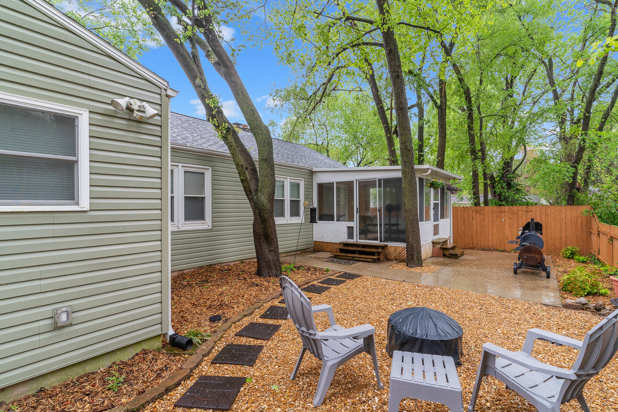 Backyard patio area with chairs, small grill, and trees behind a wooden fence.